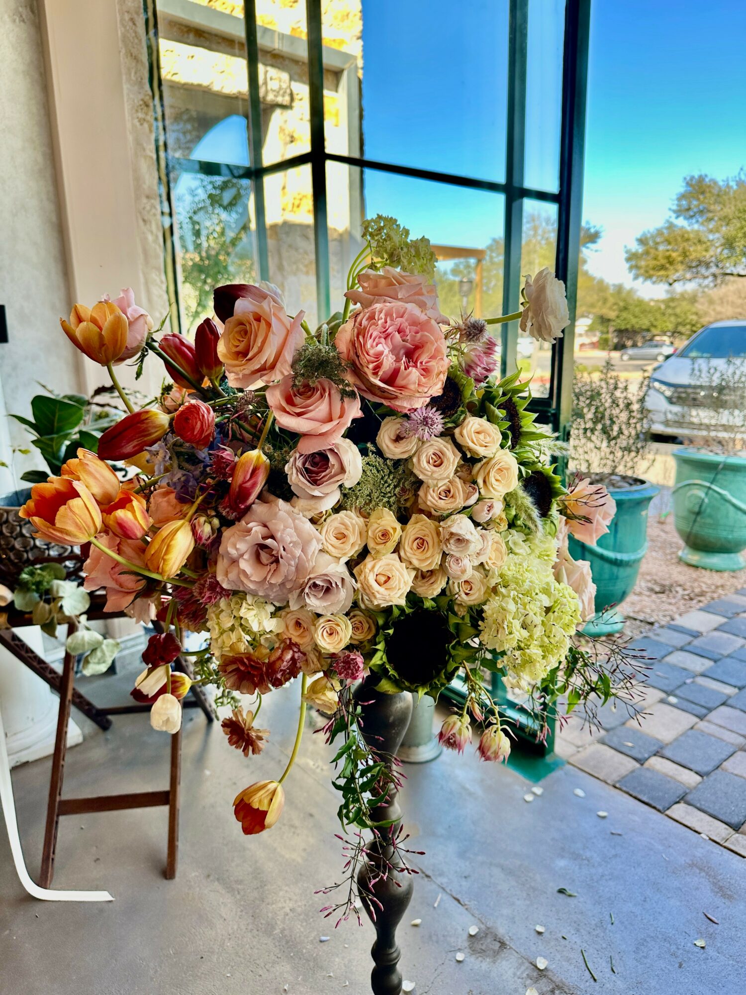 Large floral arrangement with pink, white, and cream flowers in a tall vase inside a bright room with large windows, outdoor view visible.