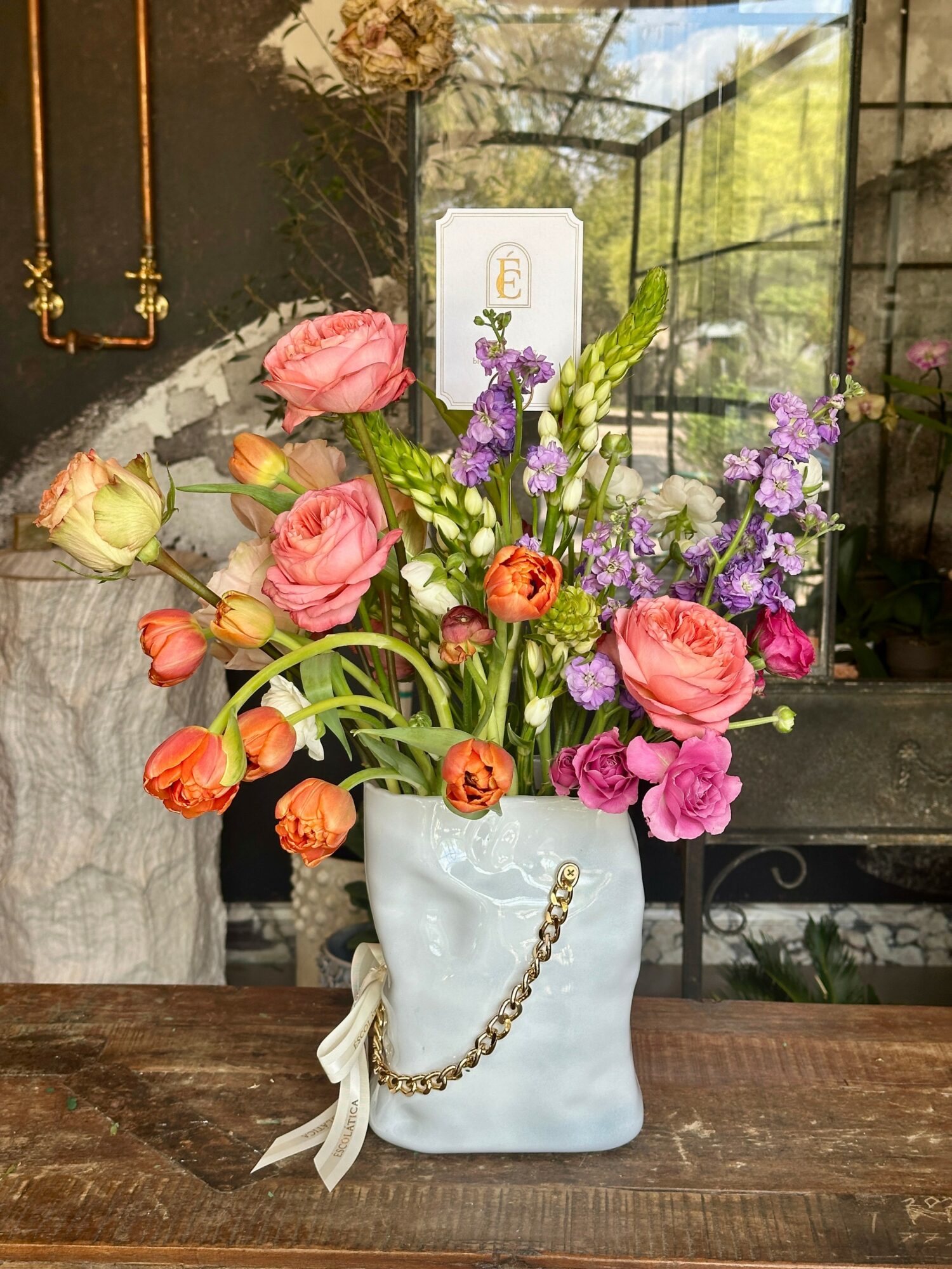 Colorful mixed flowers in a white ceramic vase on a wooden table, with a window and greenery in the background.