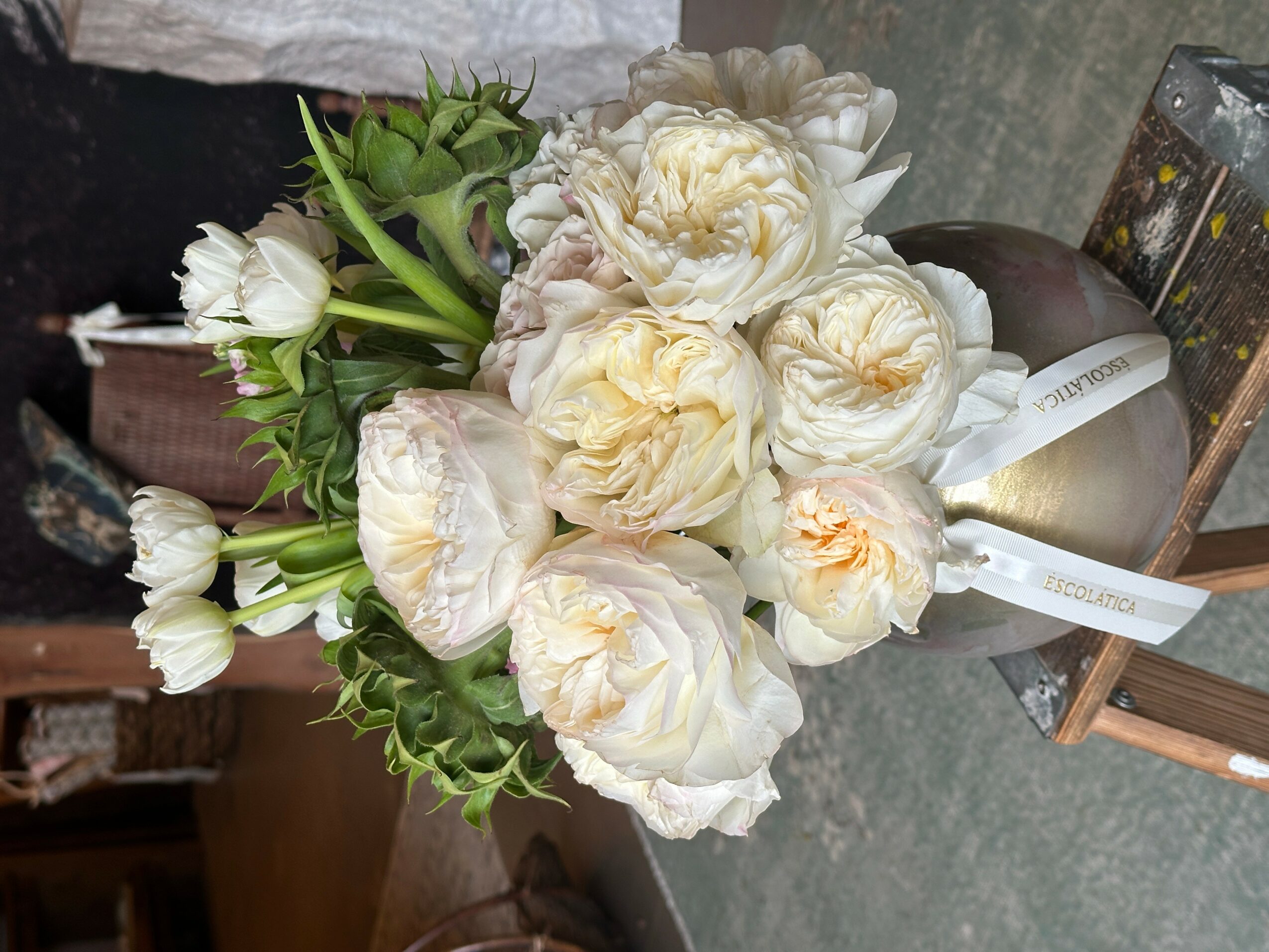 Bouquet of white and cream-colored flowers with green leaves, placed on a surface with a ribbon tied around it.