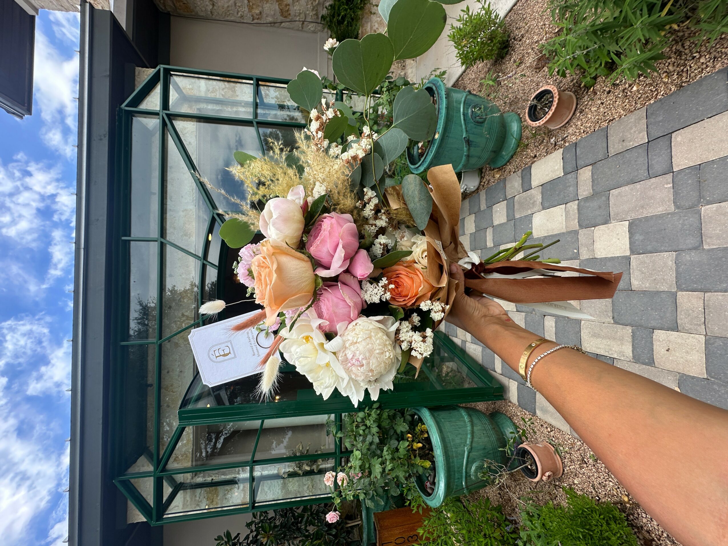 Person holding a bouquet of pink, white, and peach flowers outside a glass greenhouse, with potted plants nearby.