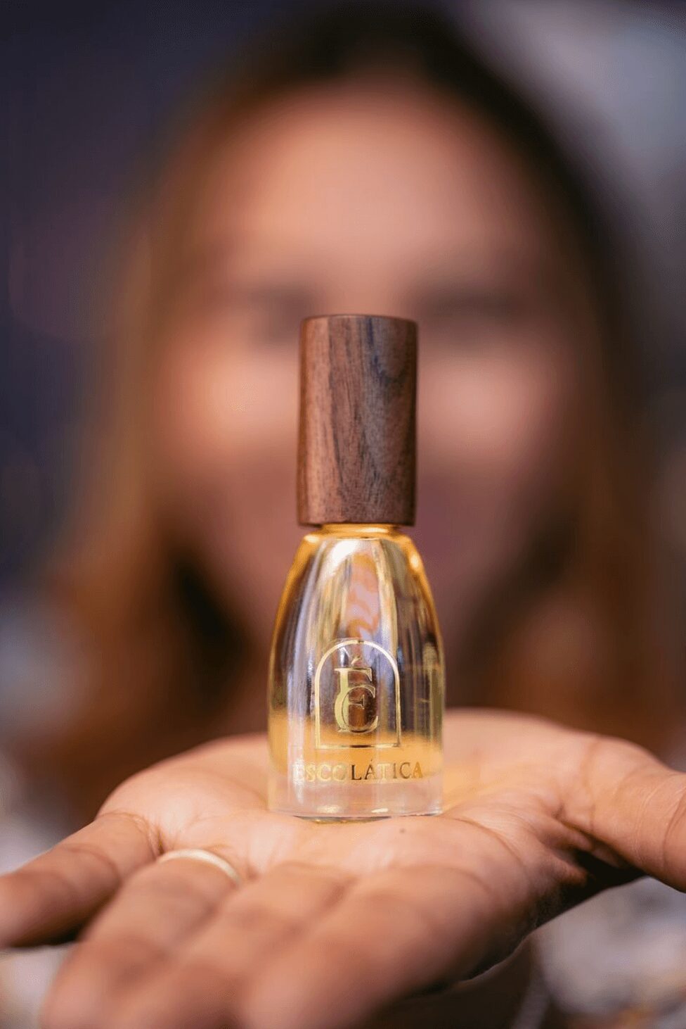 Person holding a small glass bottle with a wooden cap, focus on the bottle, blurred face in background.