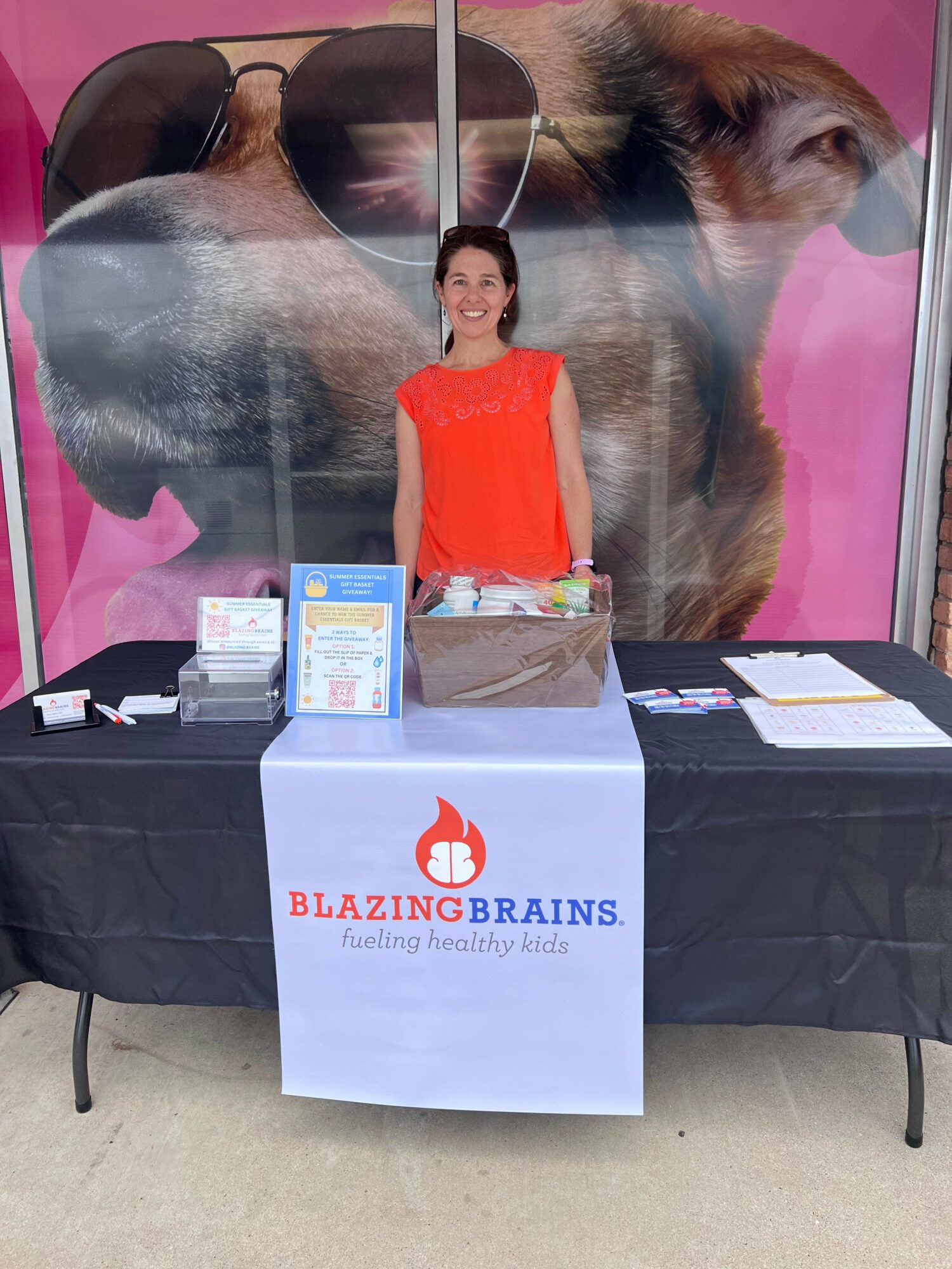 Woman standing behind a table with promotional materials, in front of a large dog poster with sunglasses, on pink background.
