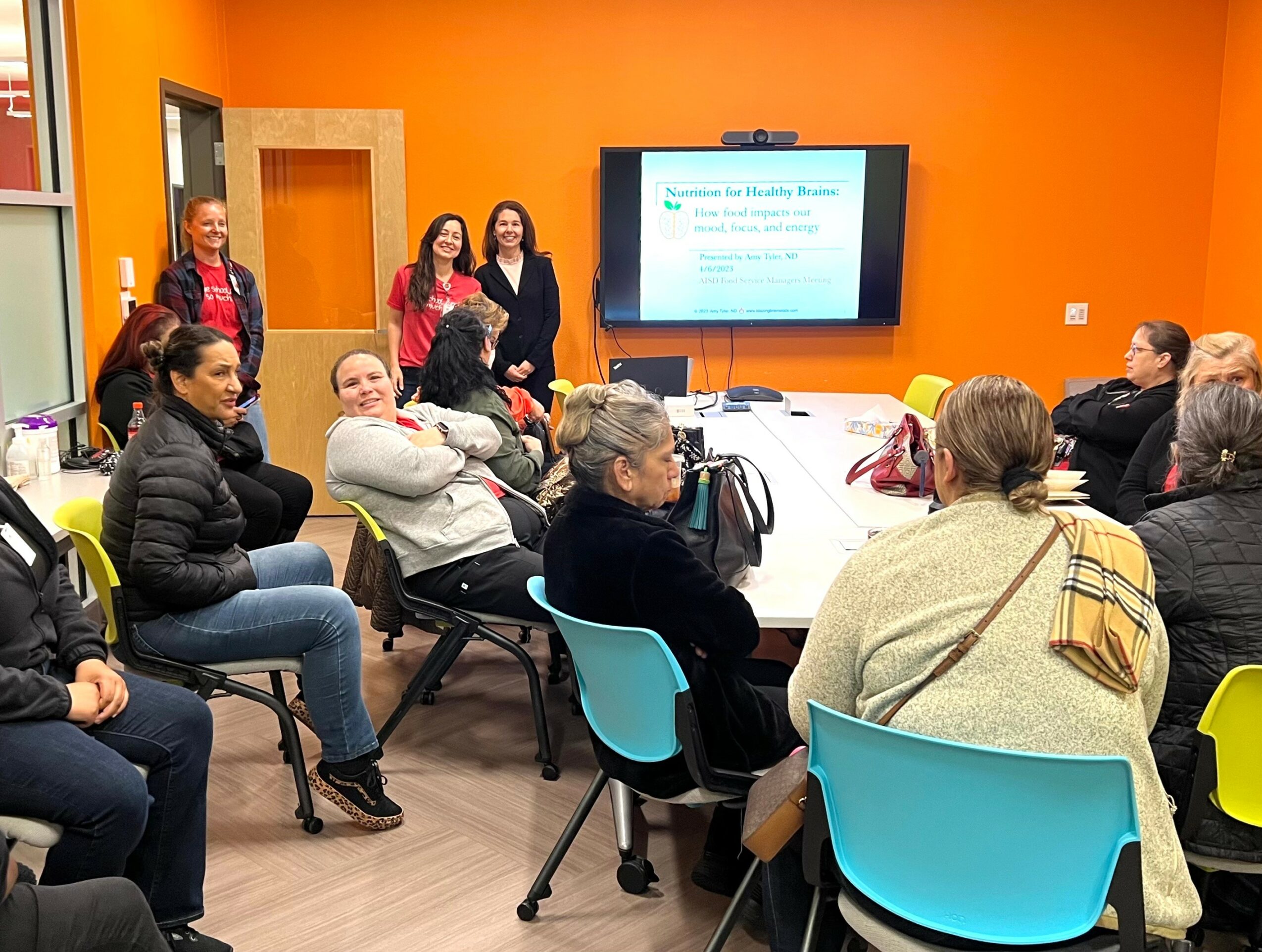 Group of people attending a presentation in a room with orange walls, seated around tables, facing a screen with a presentation slide.