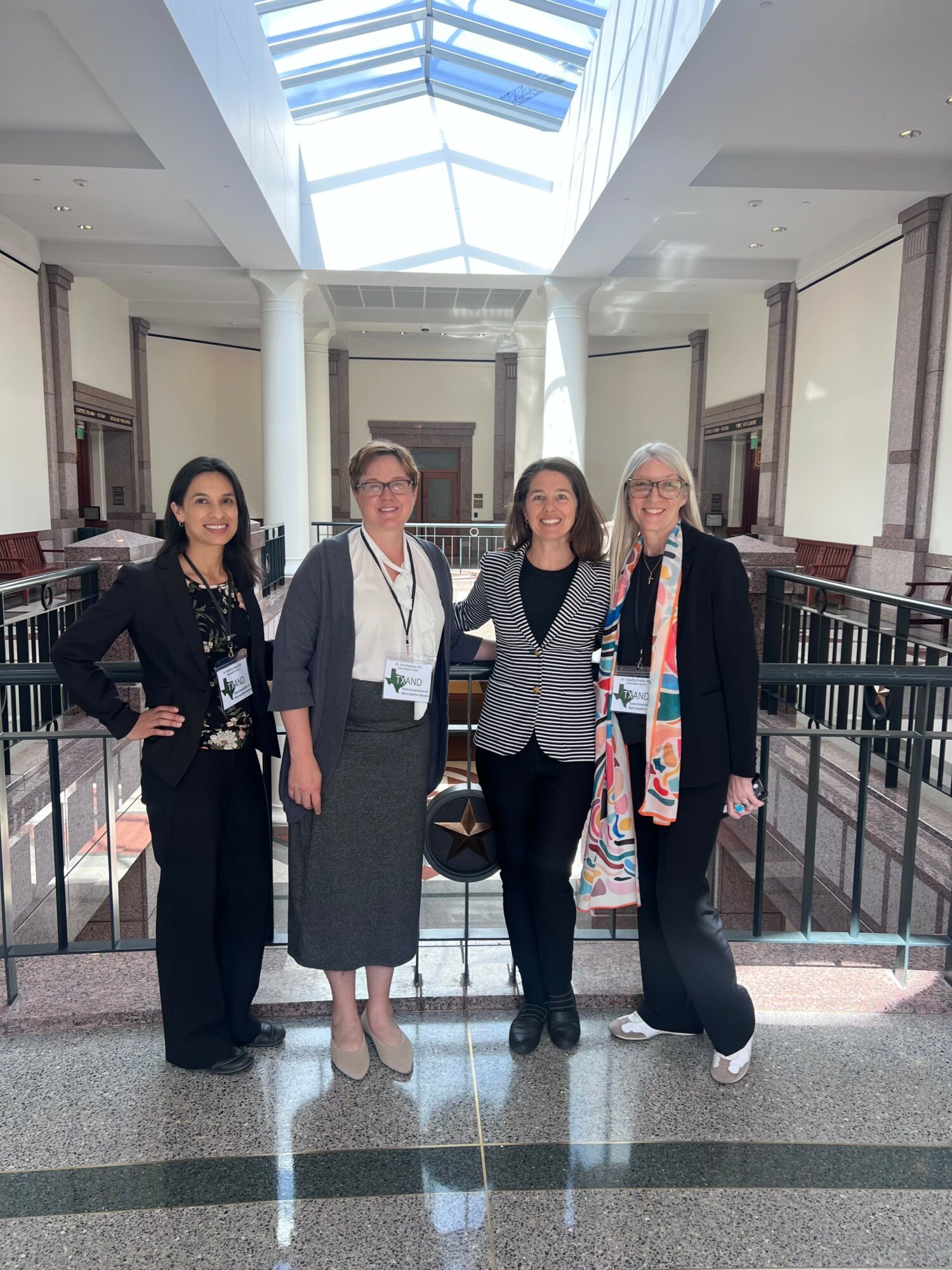 Four women standing together in a spacious, well-lit indoor area with a glass ceiling, railing, and columns.