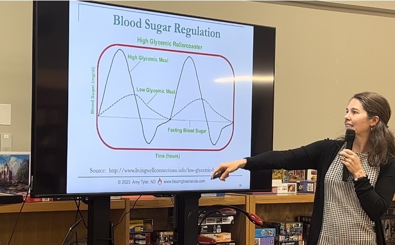 Woman presenting blood sugar regulation graph on screen, pointing at it with a microphone in hand.
