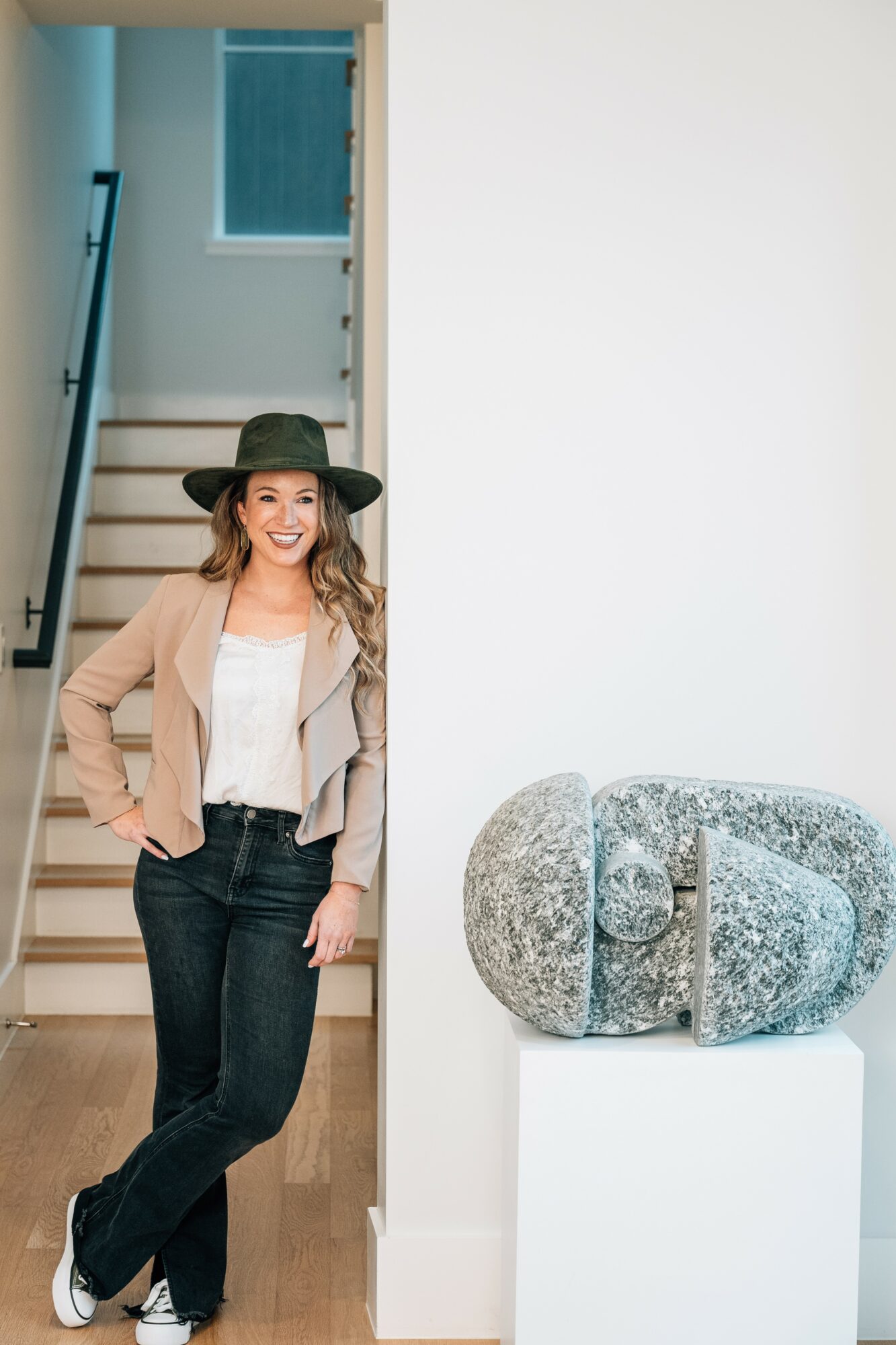 Woman with a hat standing in doorway, smiling, wearing a beige jacket and black jeans, next to stone sculpture on white pedestal.