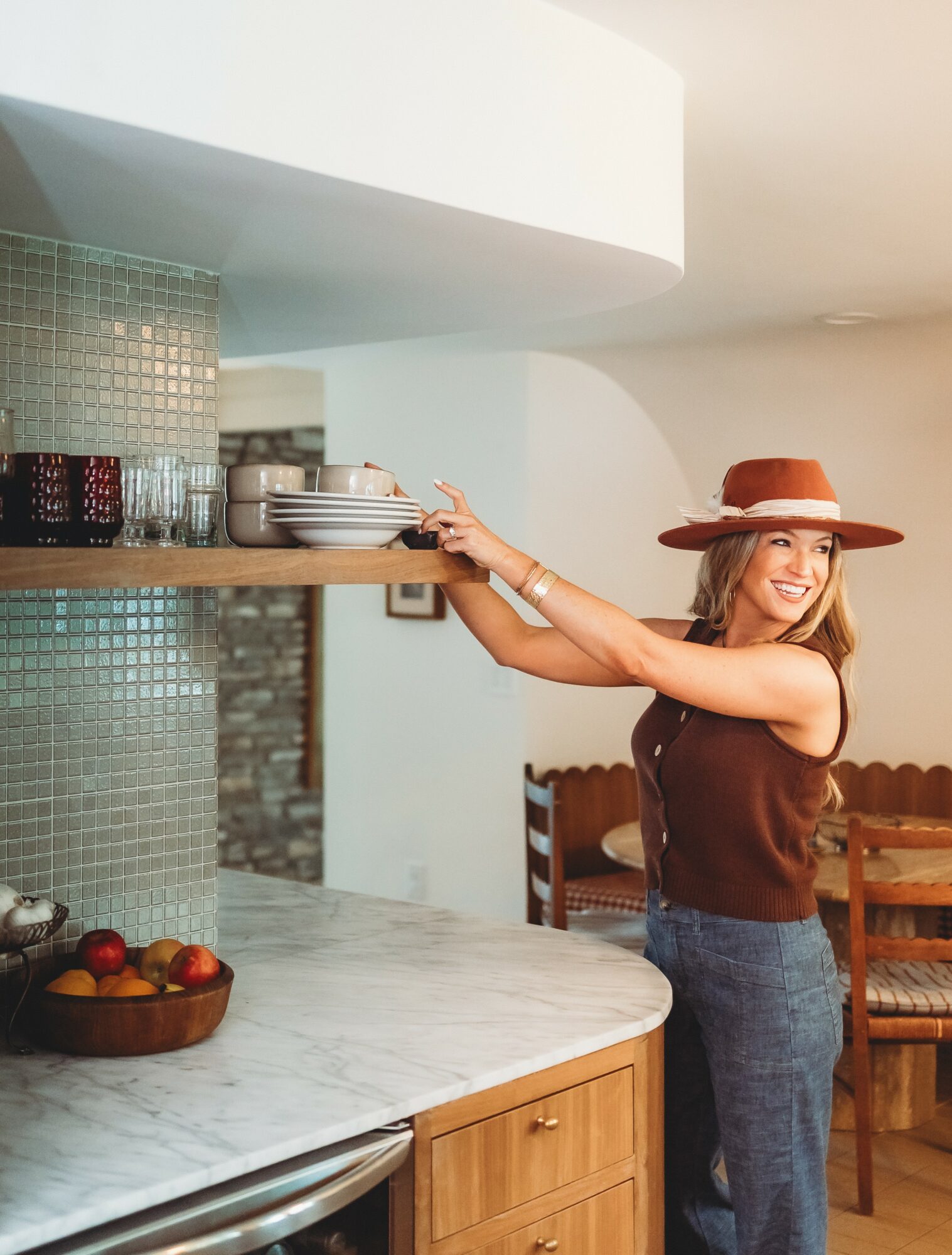 Woman in a brown top and wide-brimmed hat reaching for dishes on a kitchen shelf, smiling, with a marble countertop and wooden chairs nearby.