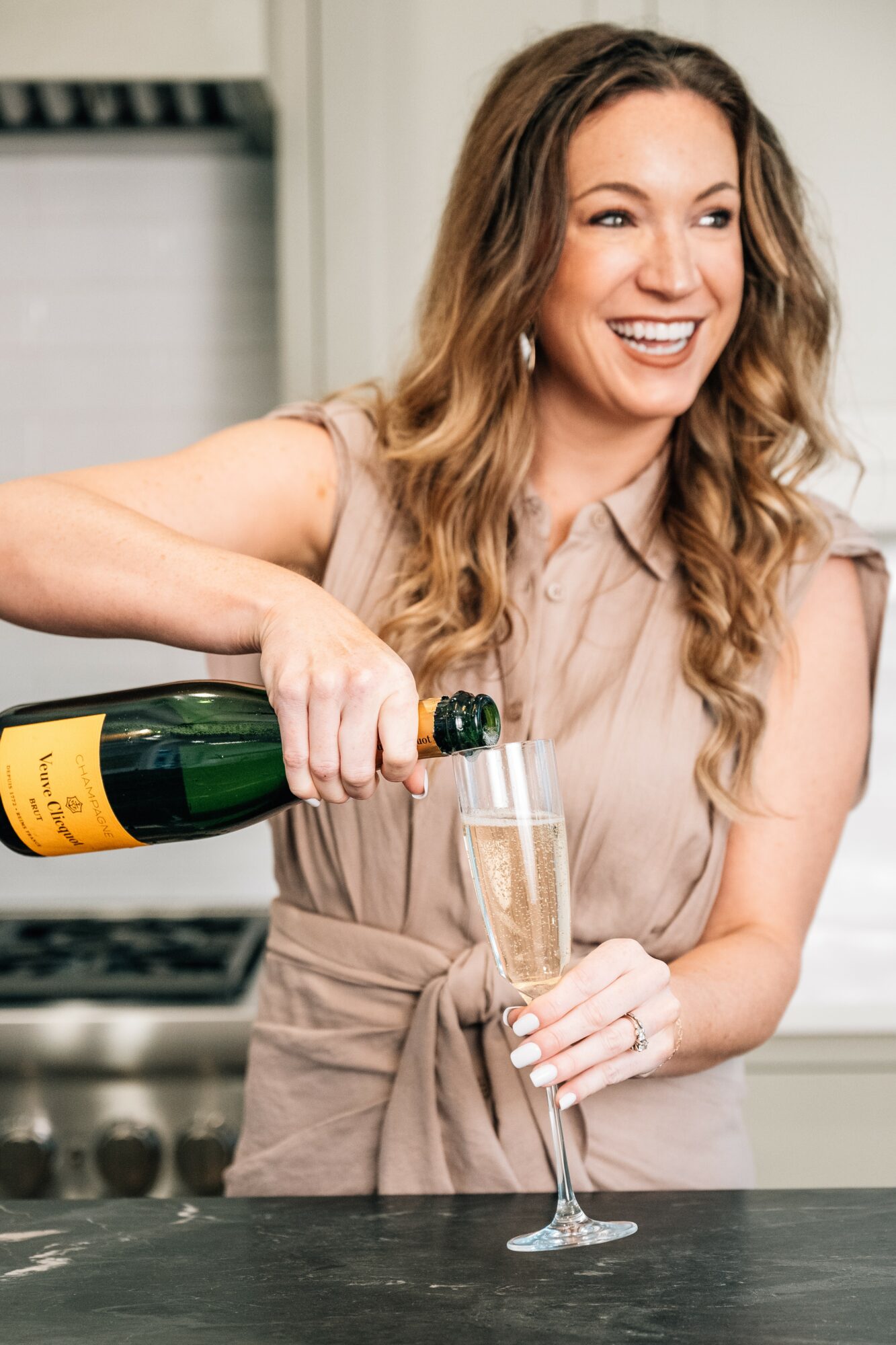 Woman pouring champagne into a glass, smiling, in a kitchen setting.