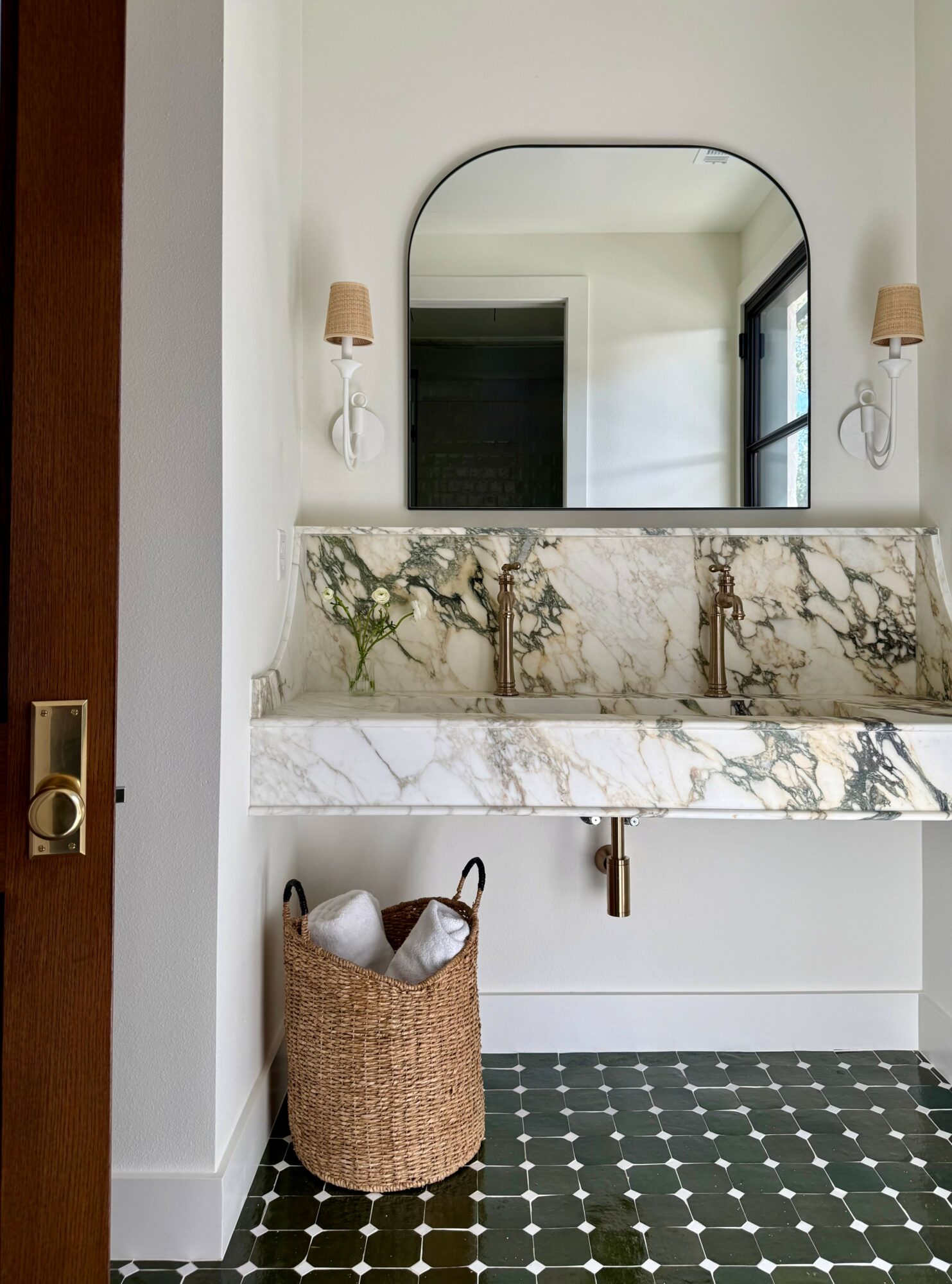 Bathroom with a marble sink, mirror, wall sconces, and a woven basket on a black and white tiled floor.