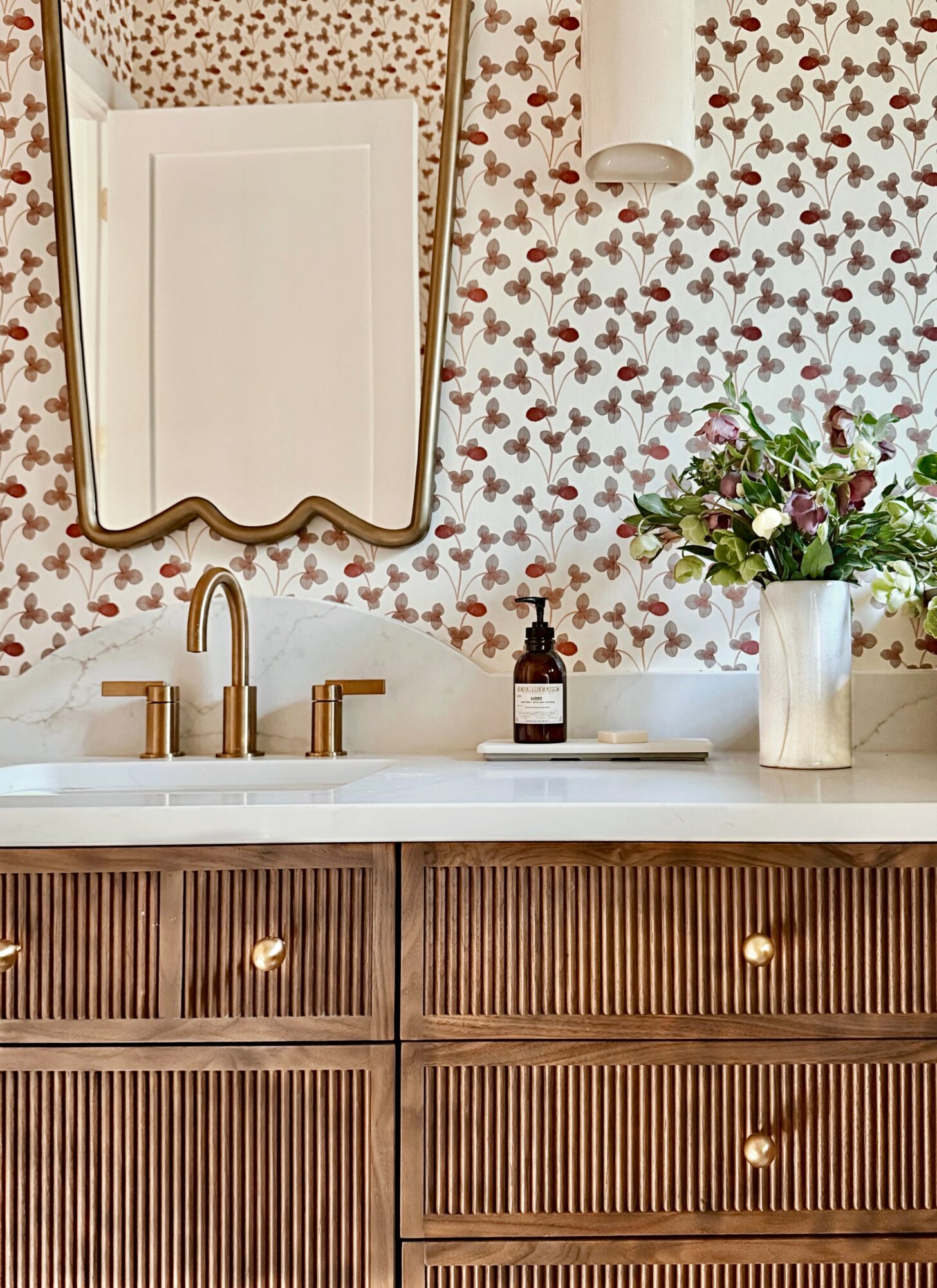 Bathroom vanity with a white countertop, gold faucet, floral wallpaper, mirror, and a vase of flowers.