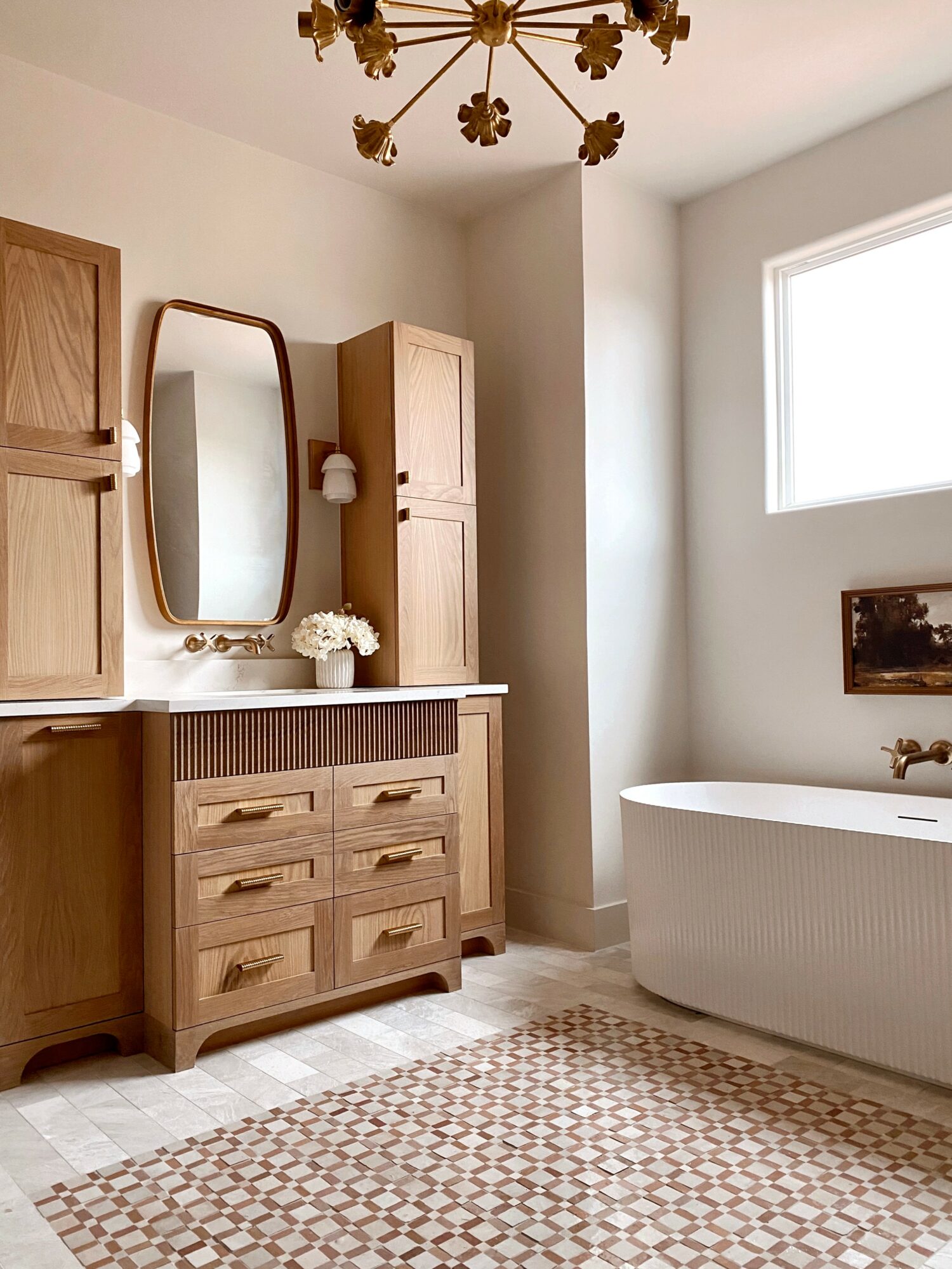 Bathroom with wooden cabinets, a mirror, a freestanding bathtub, and a window, illuminated by natural light.