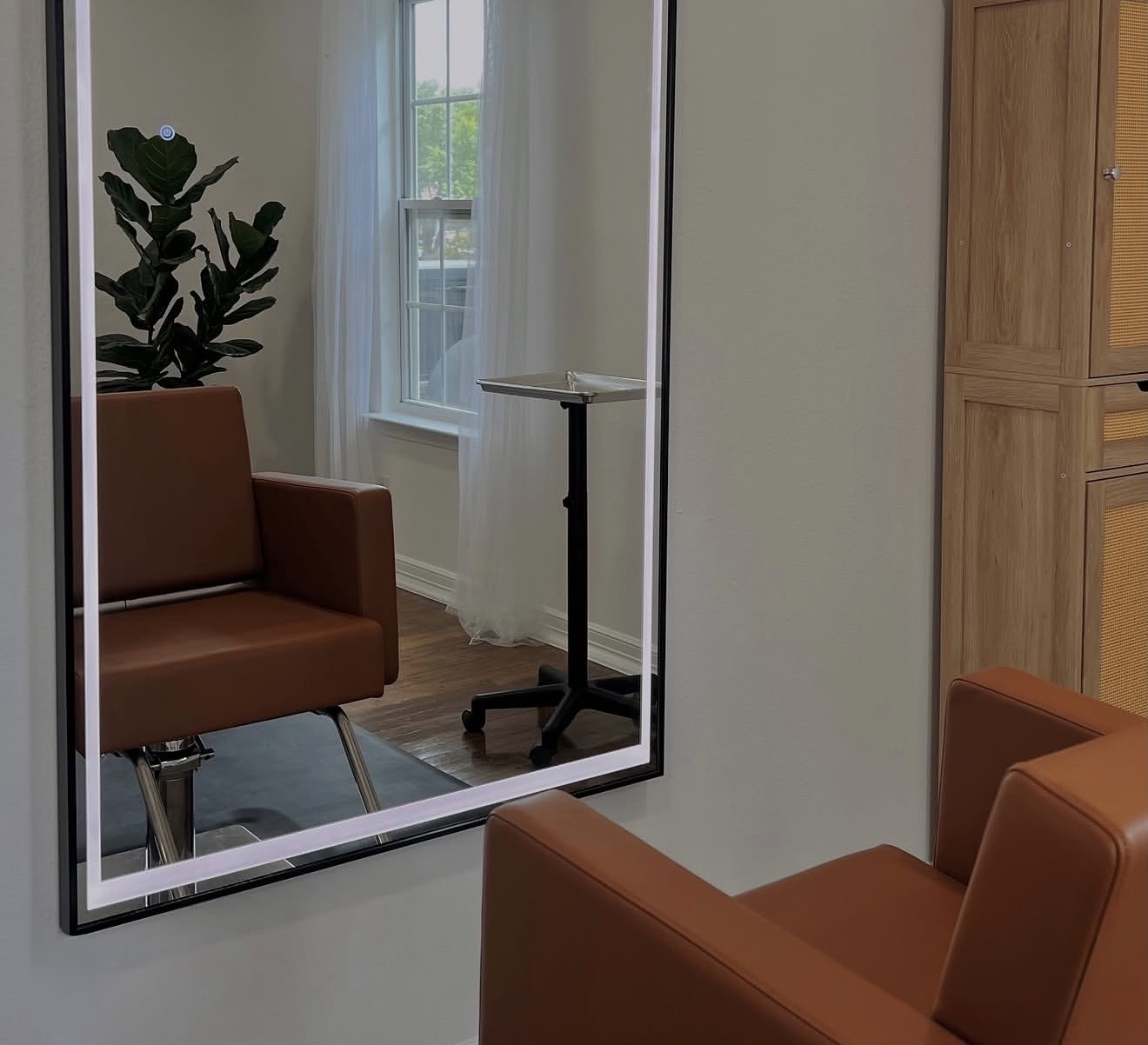 Reflection of a room with a brown chair, a window with white curtains, a tall plant, and a small table, in a mirror.