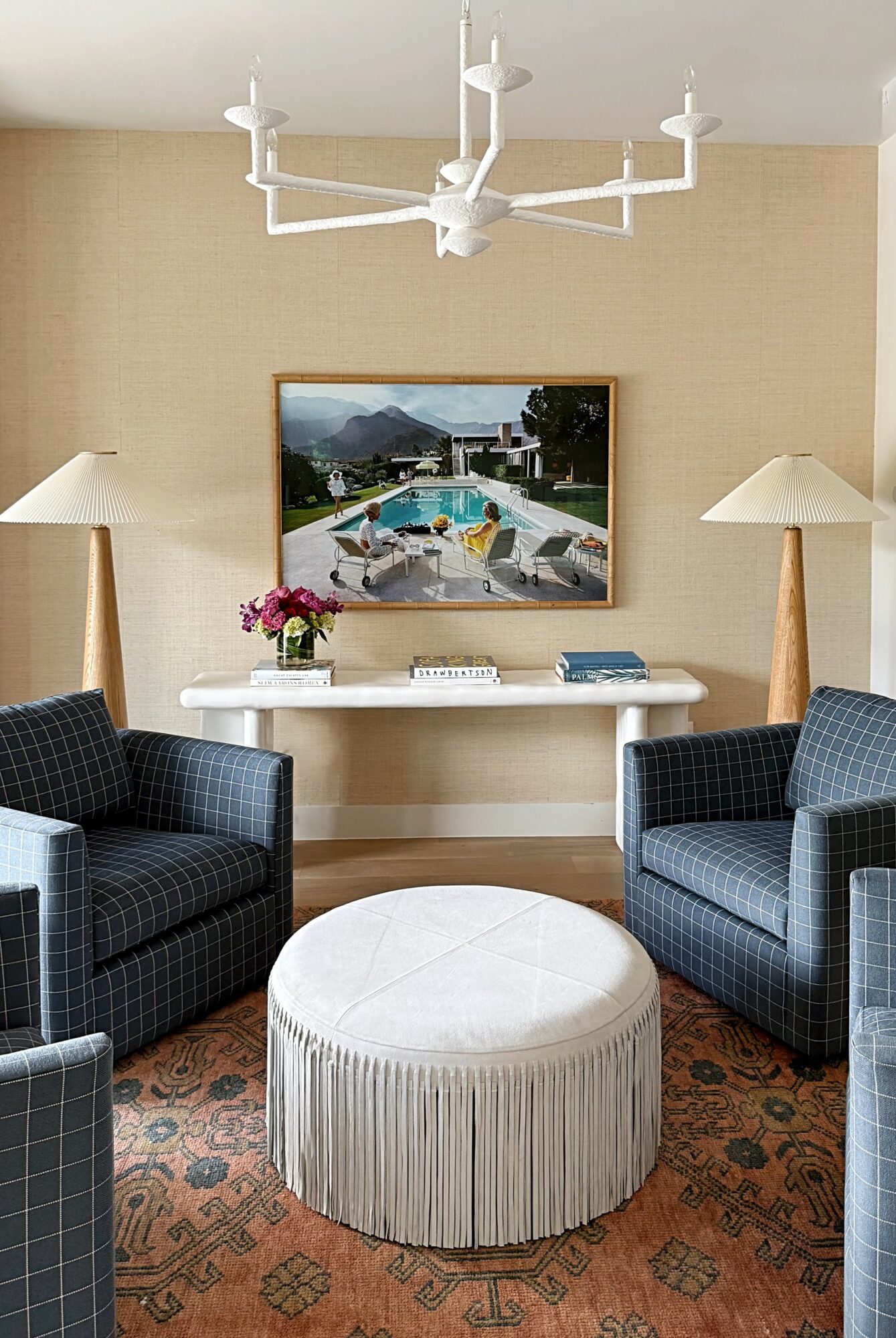 Living room with two blue armchairs, a white round ottoman, a wall-mounted TV, and two floor lamps, with a beige wall and a chandelier.