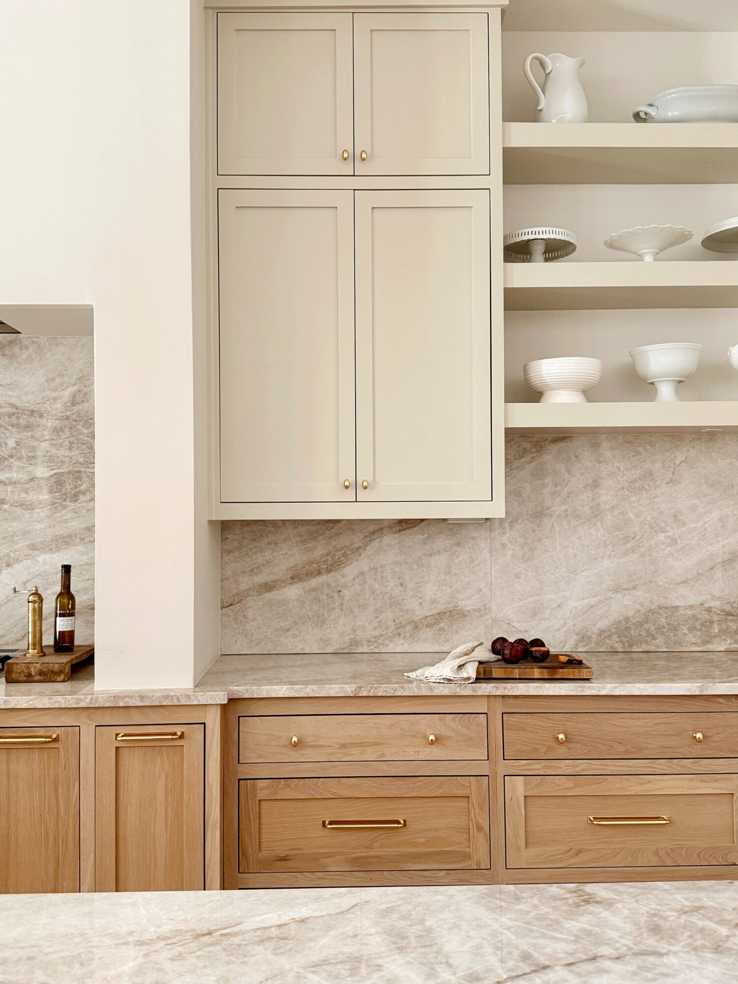 Kitchen cabinet with open shelves and drawers, beige and wood tones, with a marble countertop and some bottles and fruit.