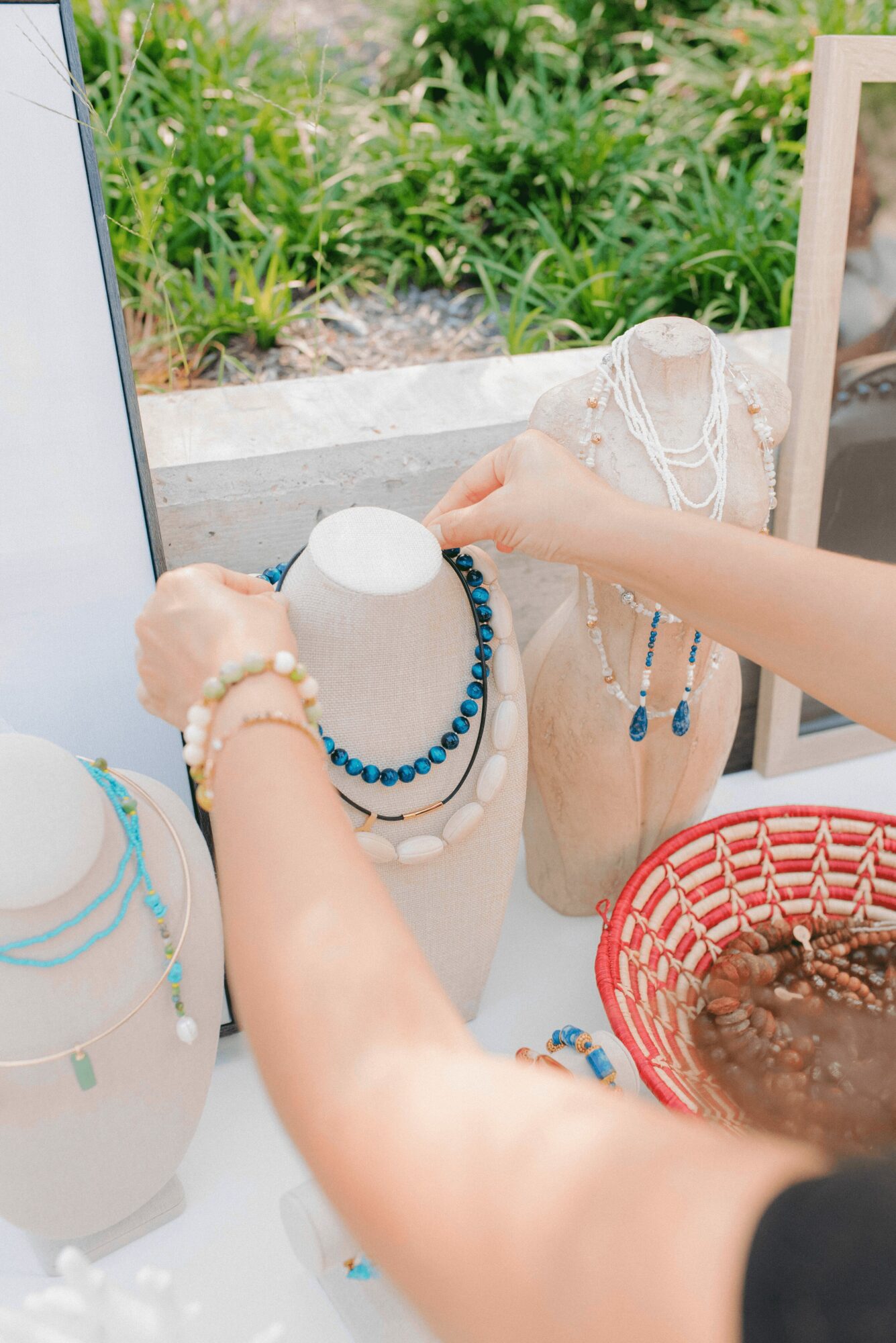 Person's hands arranging jewelry on display busts outdoors with greenery in background.