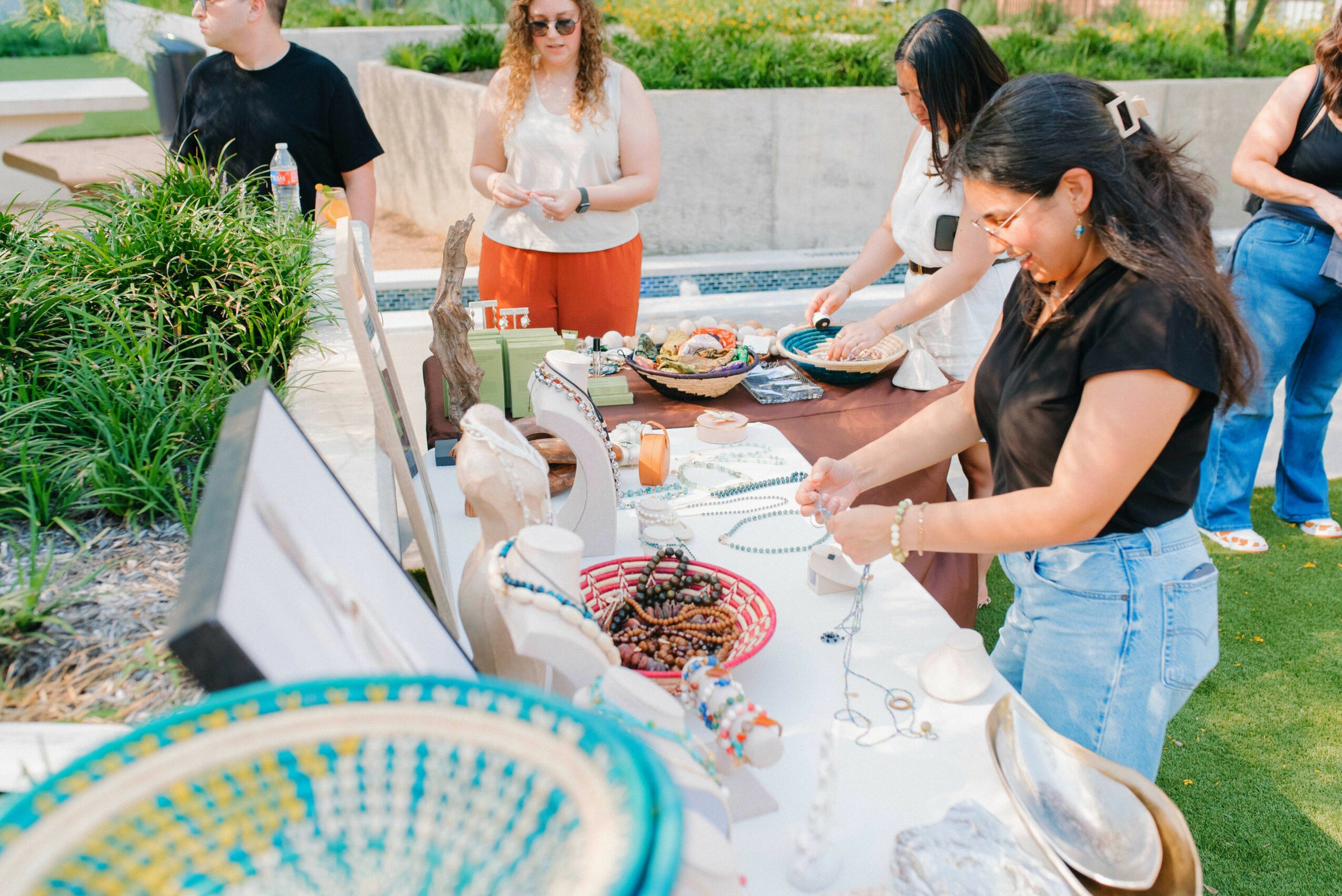Group of people at an outdoor buffet table with food and drinks, greenery in background, sunny day.