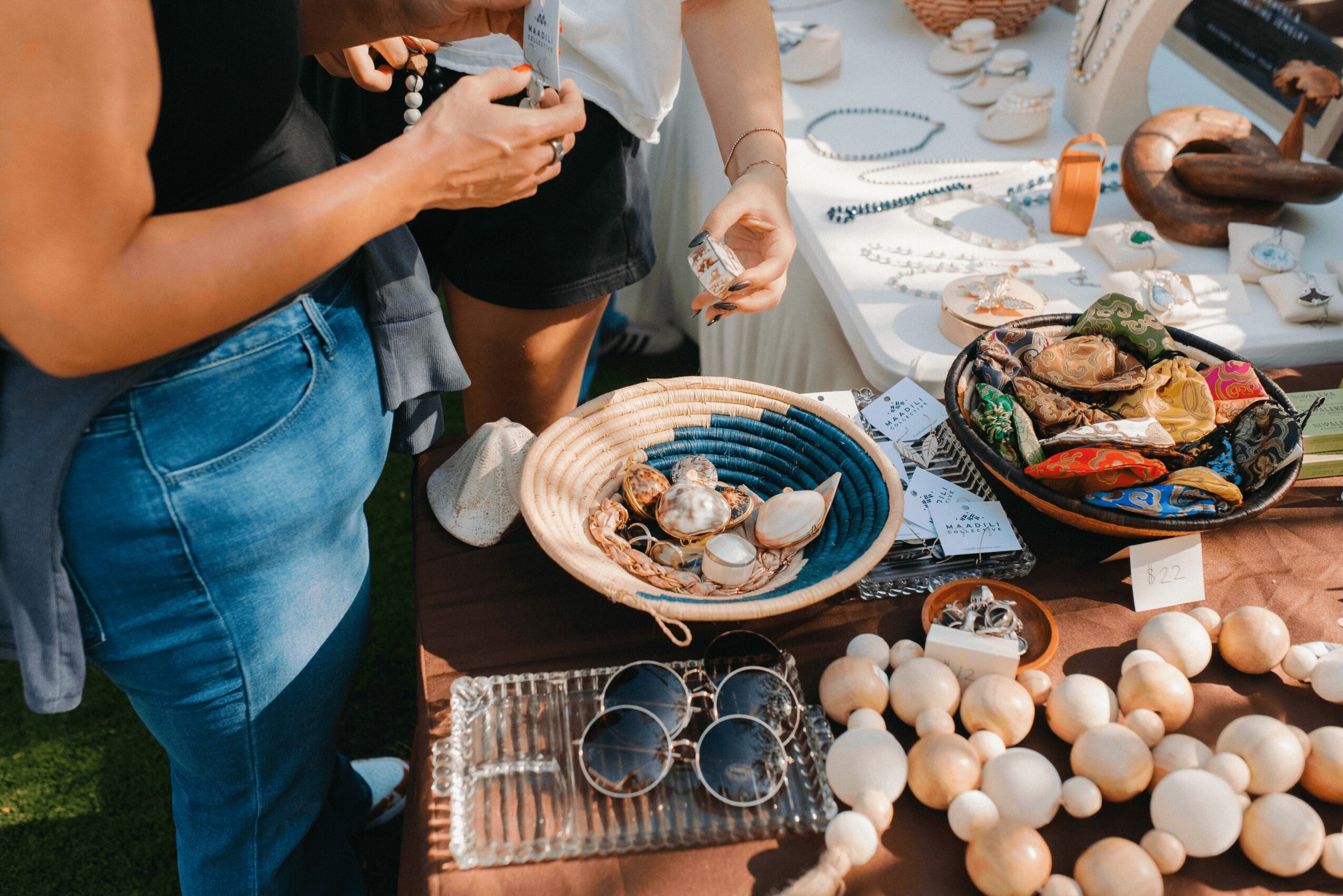 People browsing a table with jewelry, pottery, and decorative items at an outdoor market.