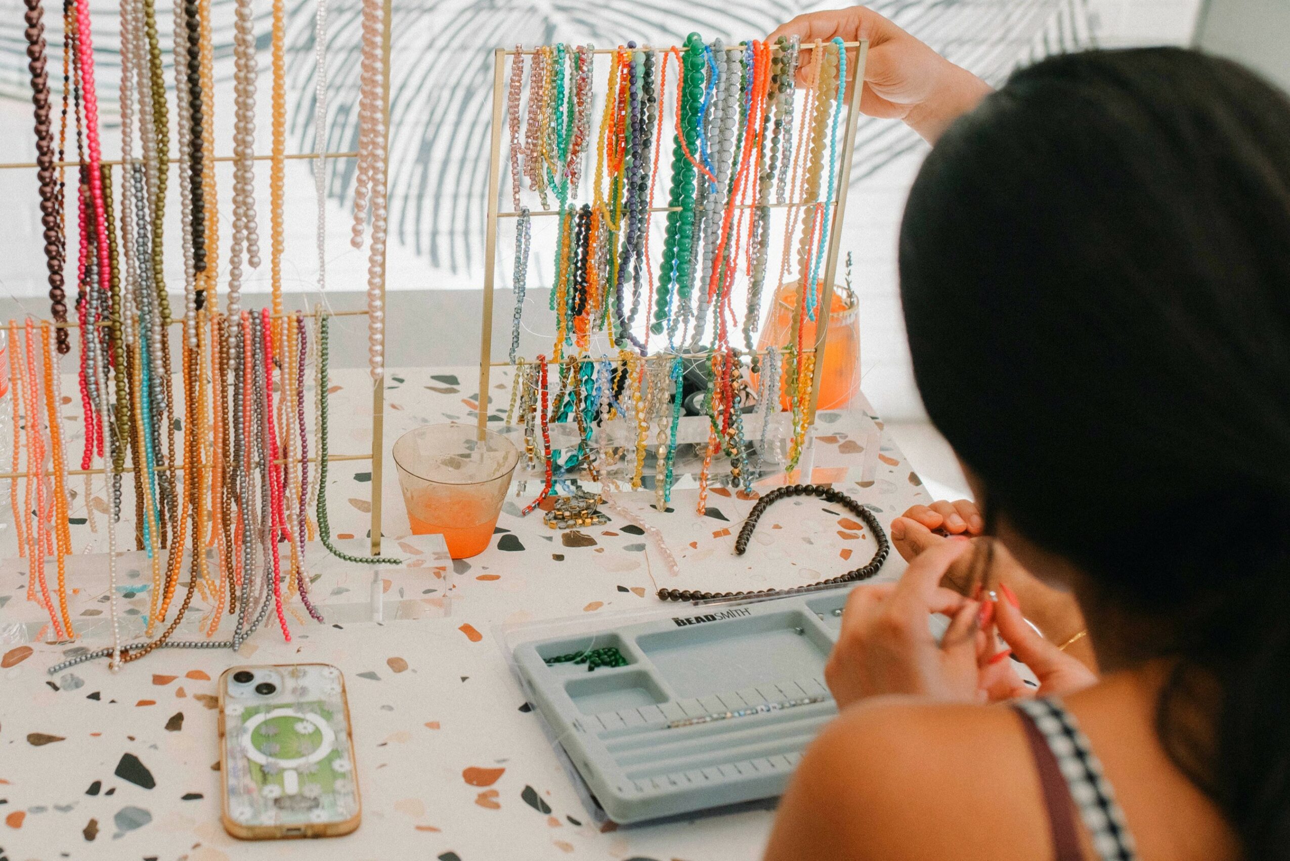 Person browsing colorful necklaces displayed on stands at a table with a tablet and a phone.