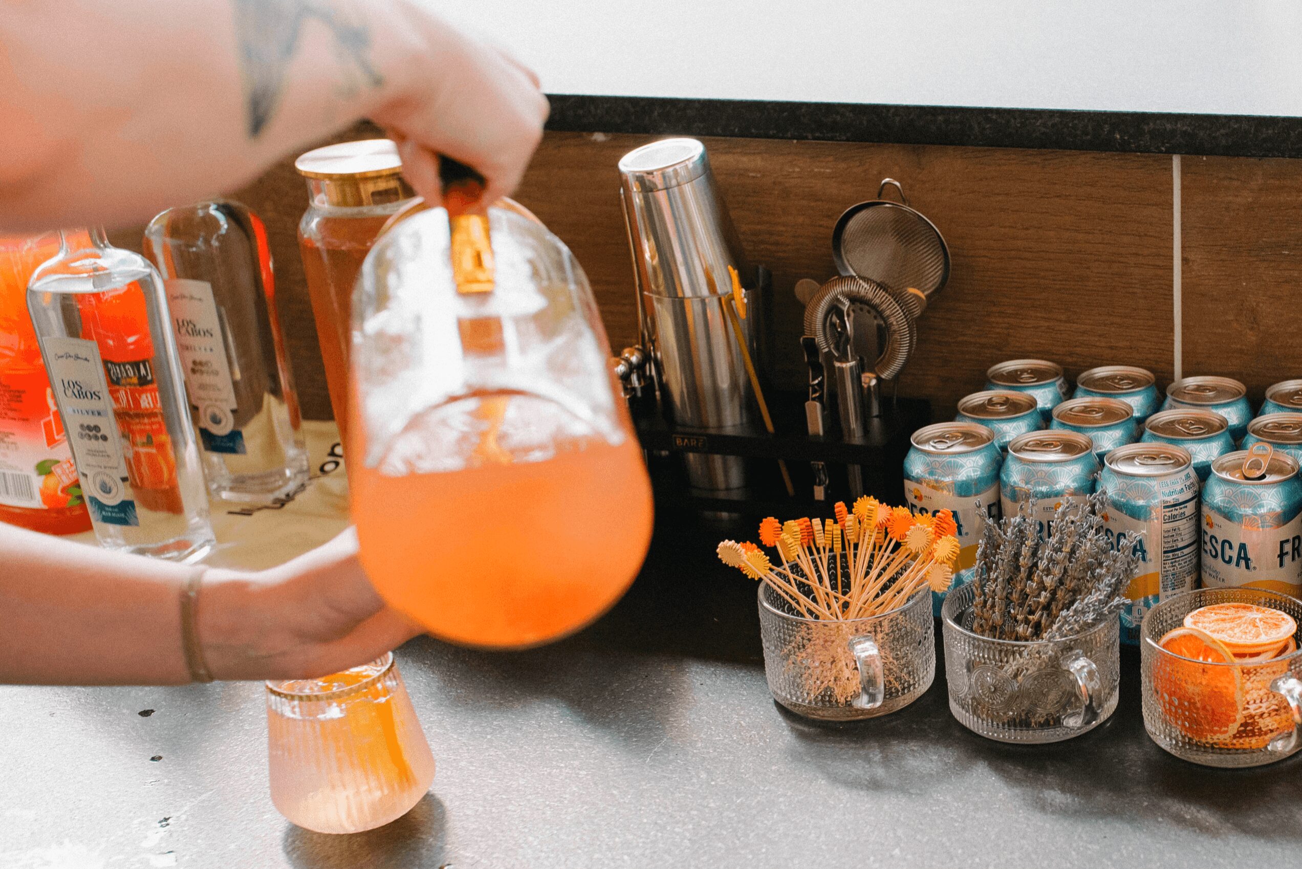 Person holding a large glass pitcher with orange-colored drink, surrounded by jars and containers on a countertop.