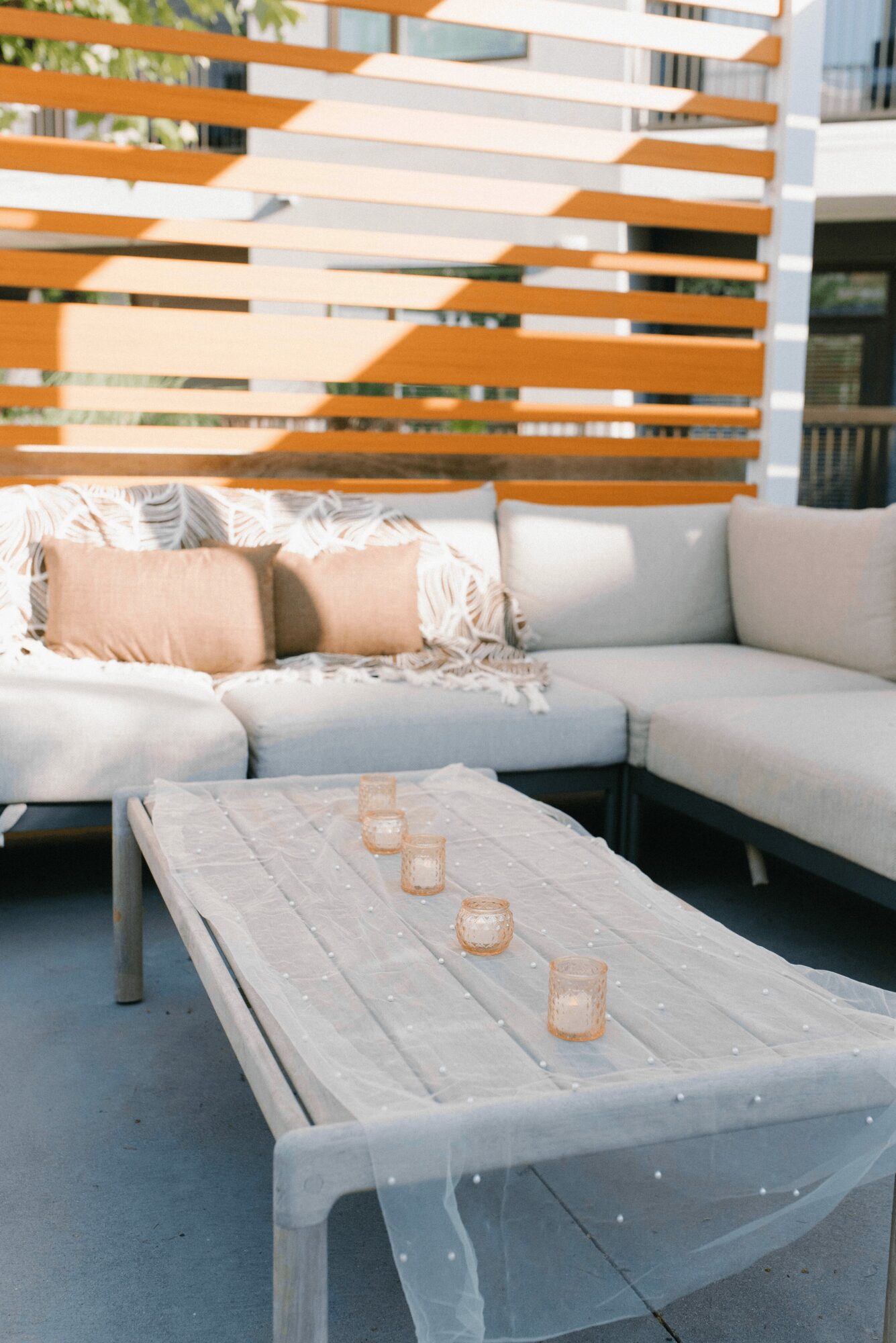 Outdoor seating area with a white sectional sofa, beige and white pillows, and a wooden table with candles.