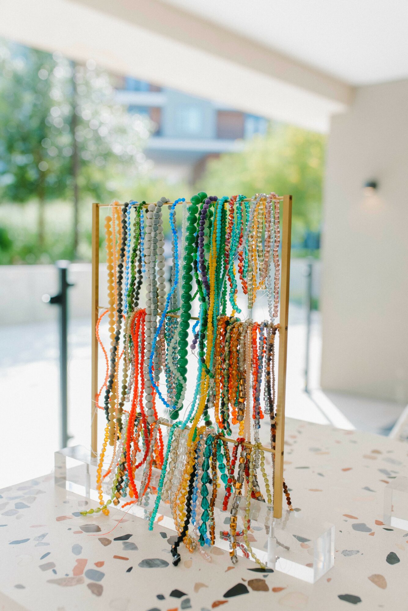 Colorful beaded necklaces hanging on a display stand indoors with a window showing trees outside