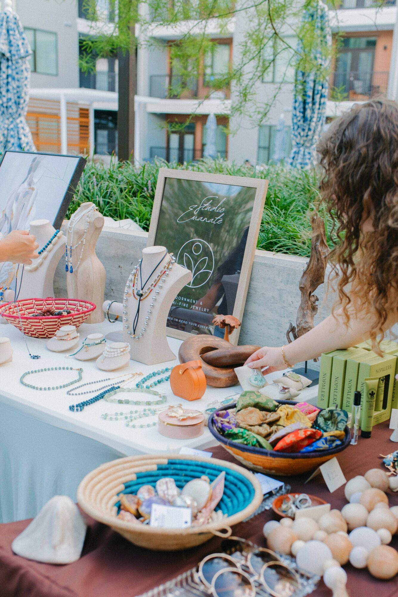People browsing jewelry and accessories at an outdoor market stall with various items on display, including necklaces, earrings, and decorative objects.
