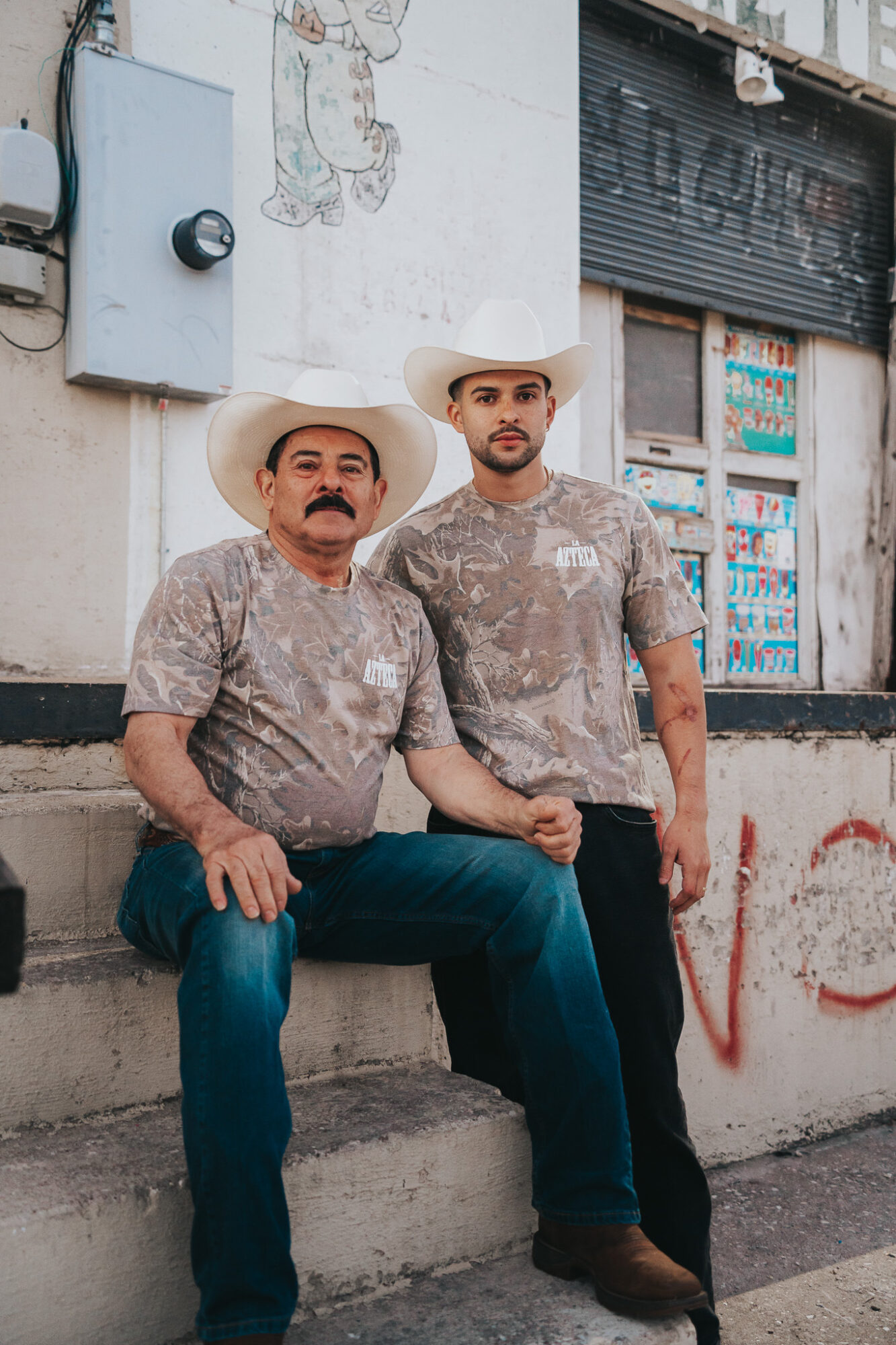 Two men wearing cowboy hats and camouflage shirts sit on a concrete ledge in front of a wall with graffiti and a closed shop. One man has a mustache and the other has a beard.