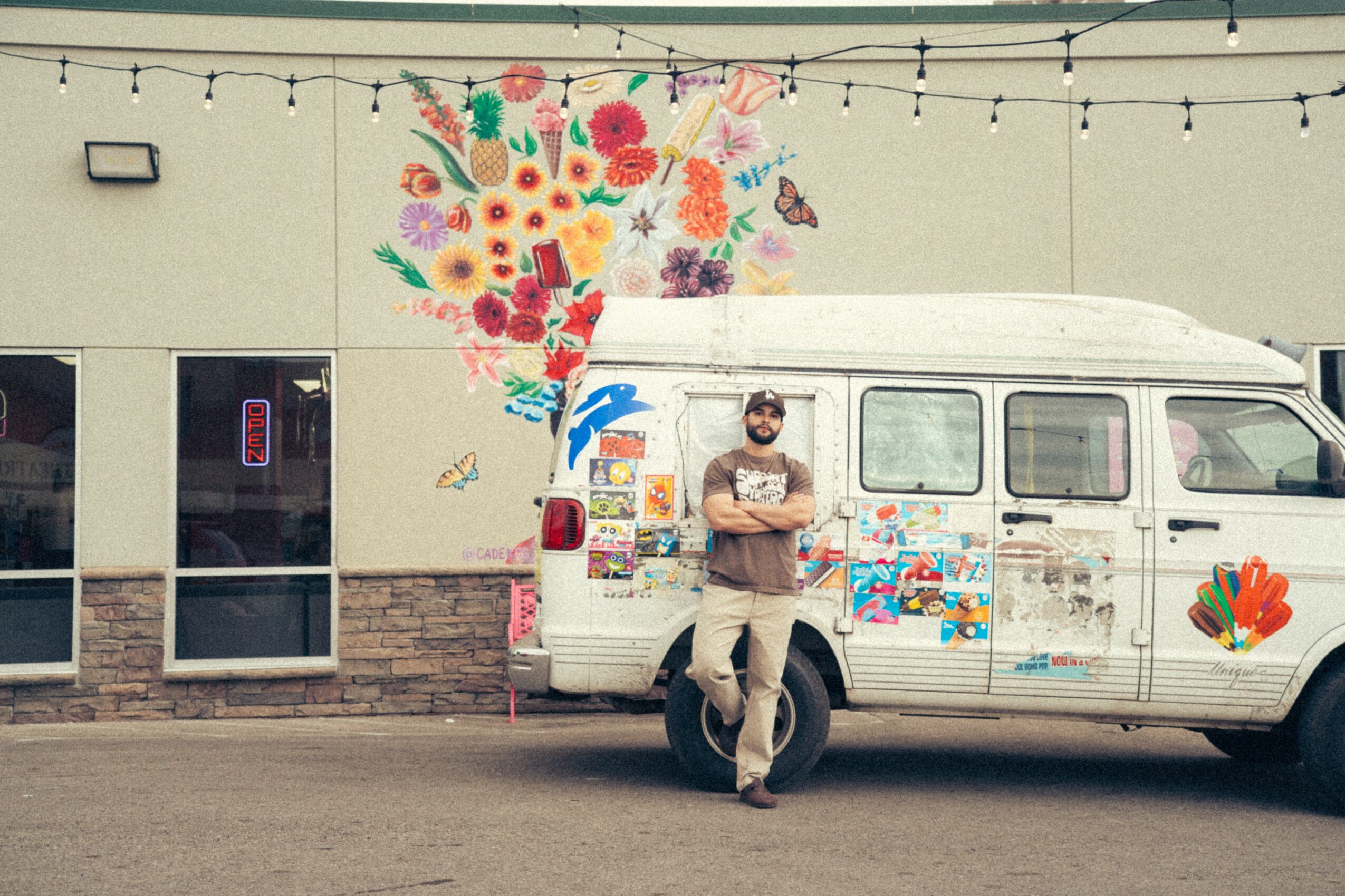 Man standing in front of a decorated van with colorful artwork, in front of a building with string lights overhead.