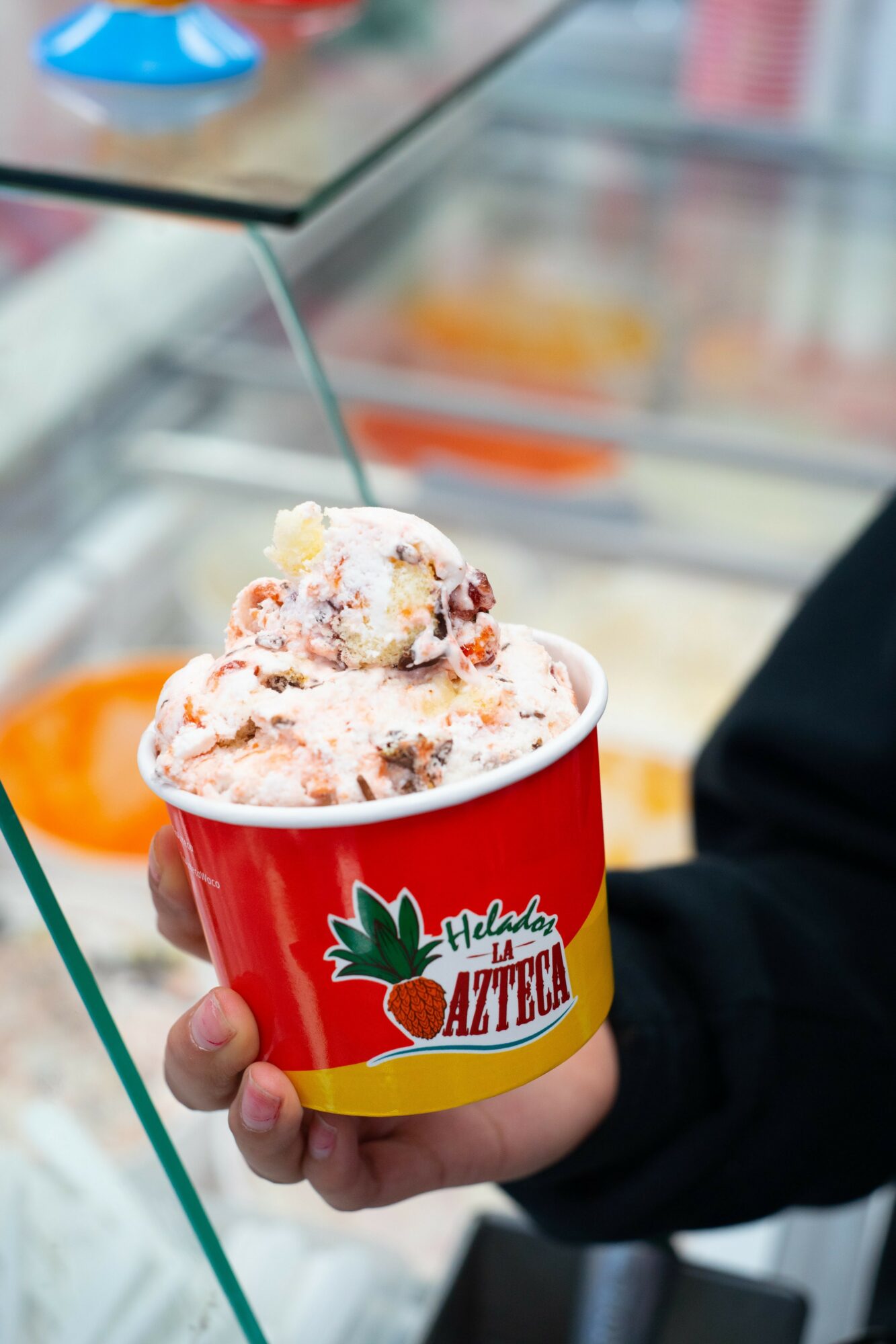 Person holding a red cup of ice cream with fruit pieces, in a store setting.