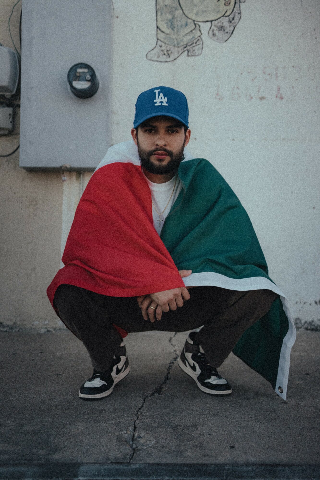 Young man crouching outdoors, wearing a blue cap, with an Italian flag draped over his shoulders, against a wall.