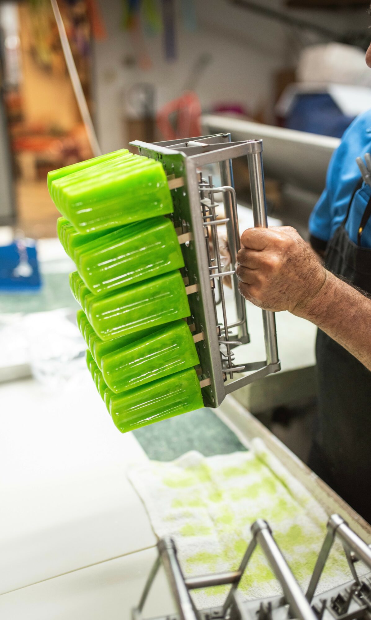 Person holding a metal rack with six green plastic containers in a kitchen or workshop setting.