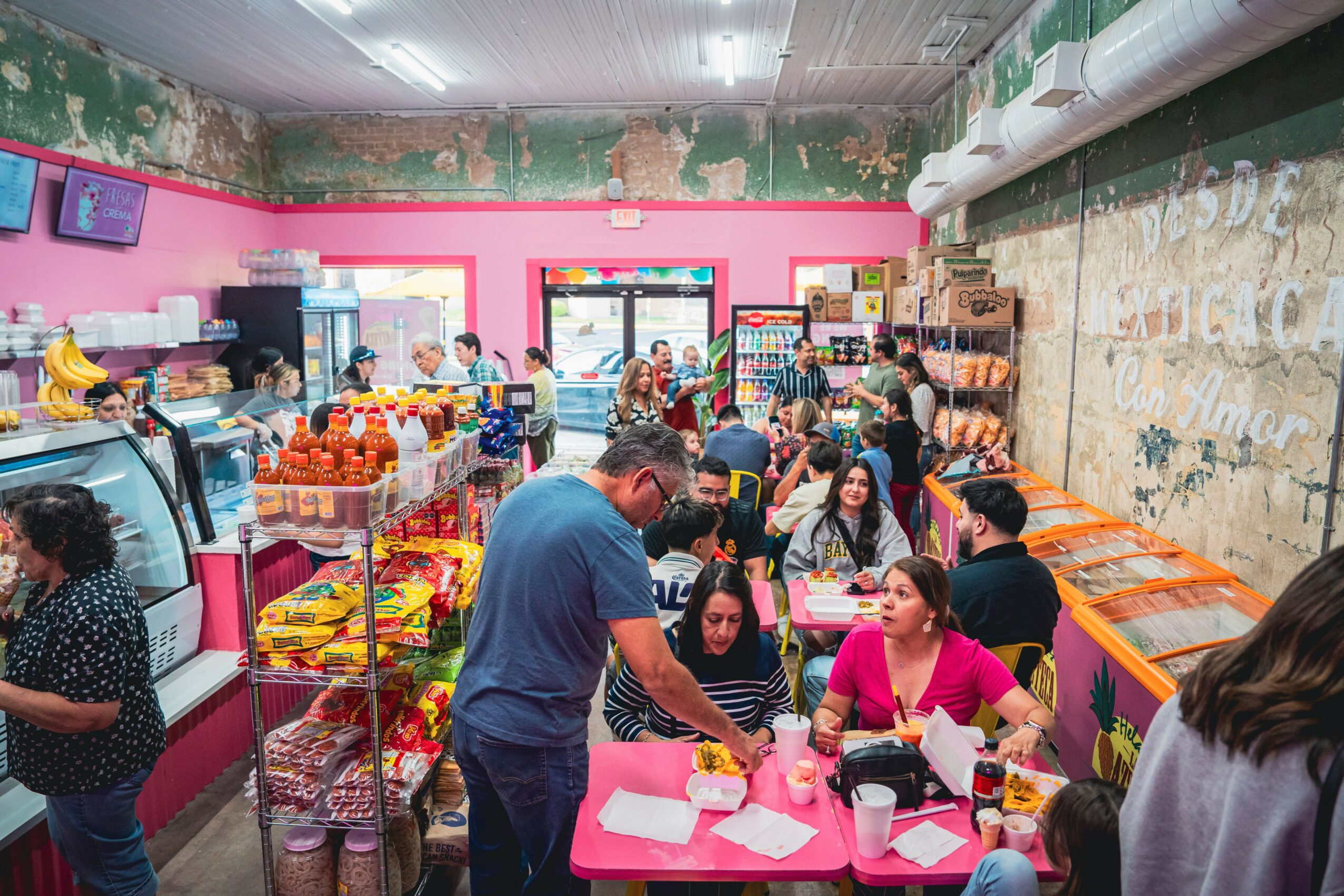 People shopping inside a colorful store with pink walls, shelves of snacks, and a busy atmosphere.