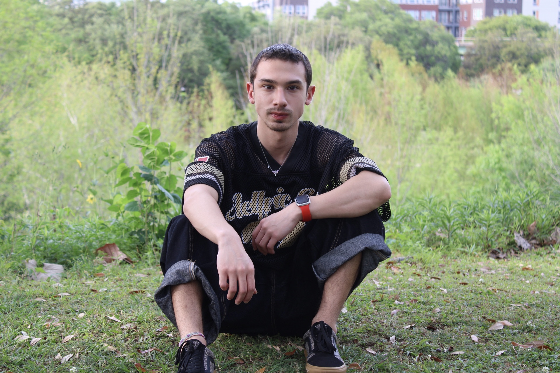 Young man sitting outdoors on grass with greenery and trees in background, wearing black clothing and sneakers, looking at camera.