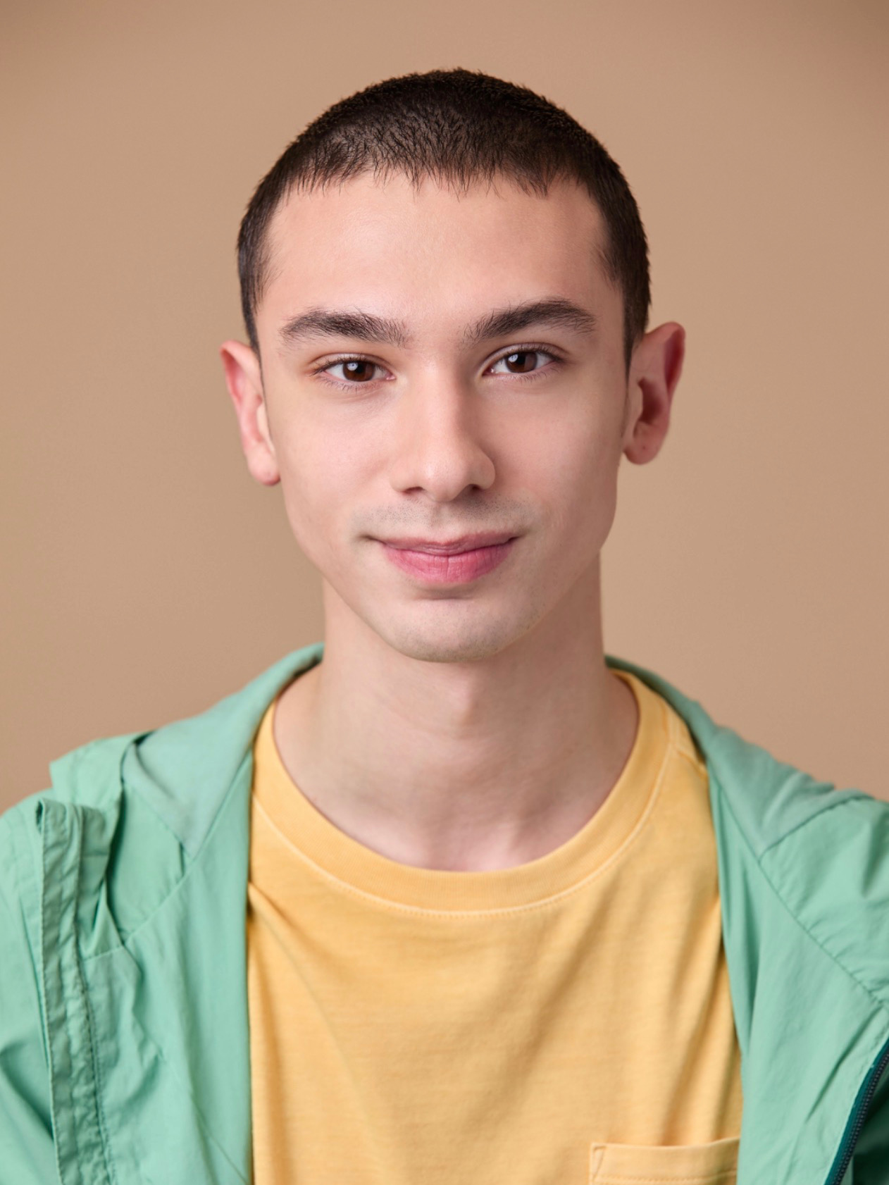 Young man with short dark hair, wearing a yellow shirt and green jacket, smiling softly against a beige background.