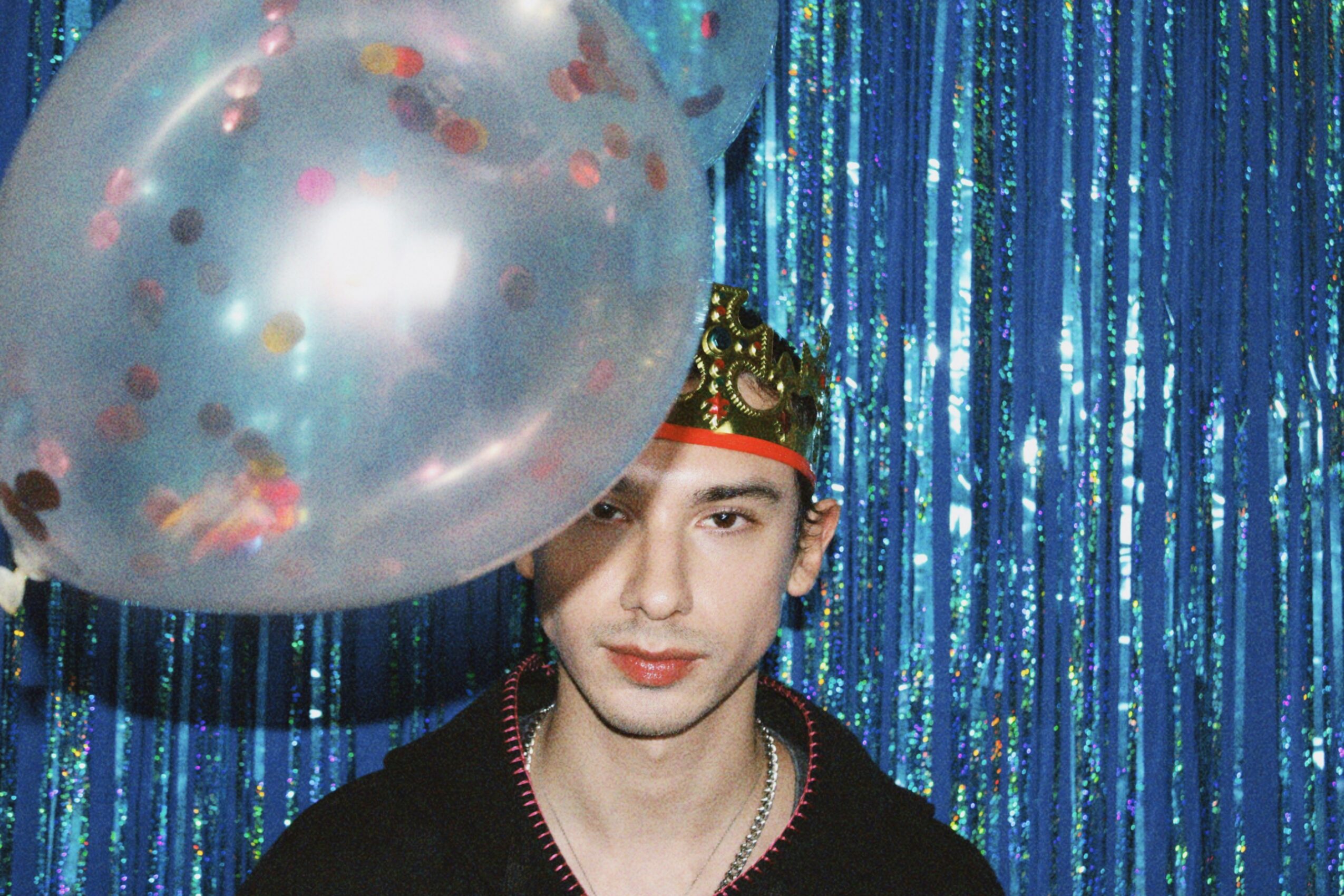 Person wearing a crown and black clothing, standing in front of a shiny blue curtain, holding a transparent balloon with confetti.
