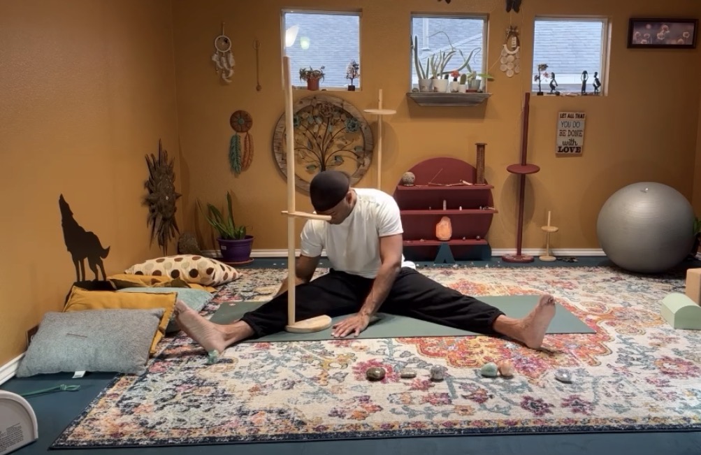 Person sitting on a colorful rug in a room with windows, plants, and various decorations, stretching legs wide apart.