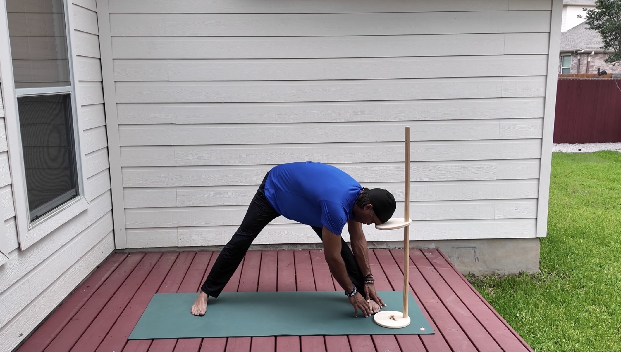 Person in blue shirt bending forward on a yoga mat outdoors, with a stick nearby, on a wooden deck.