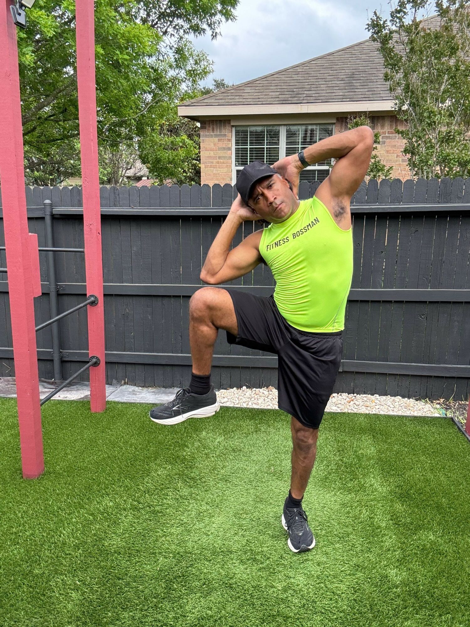 Athlete in neon green shirt and black shorts stretching outdoors on green grass, with a black fence and trees in background.