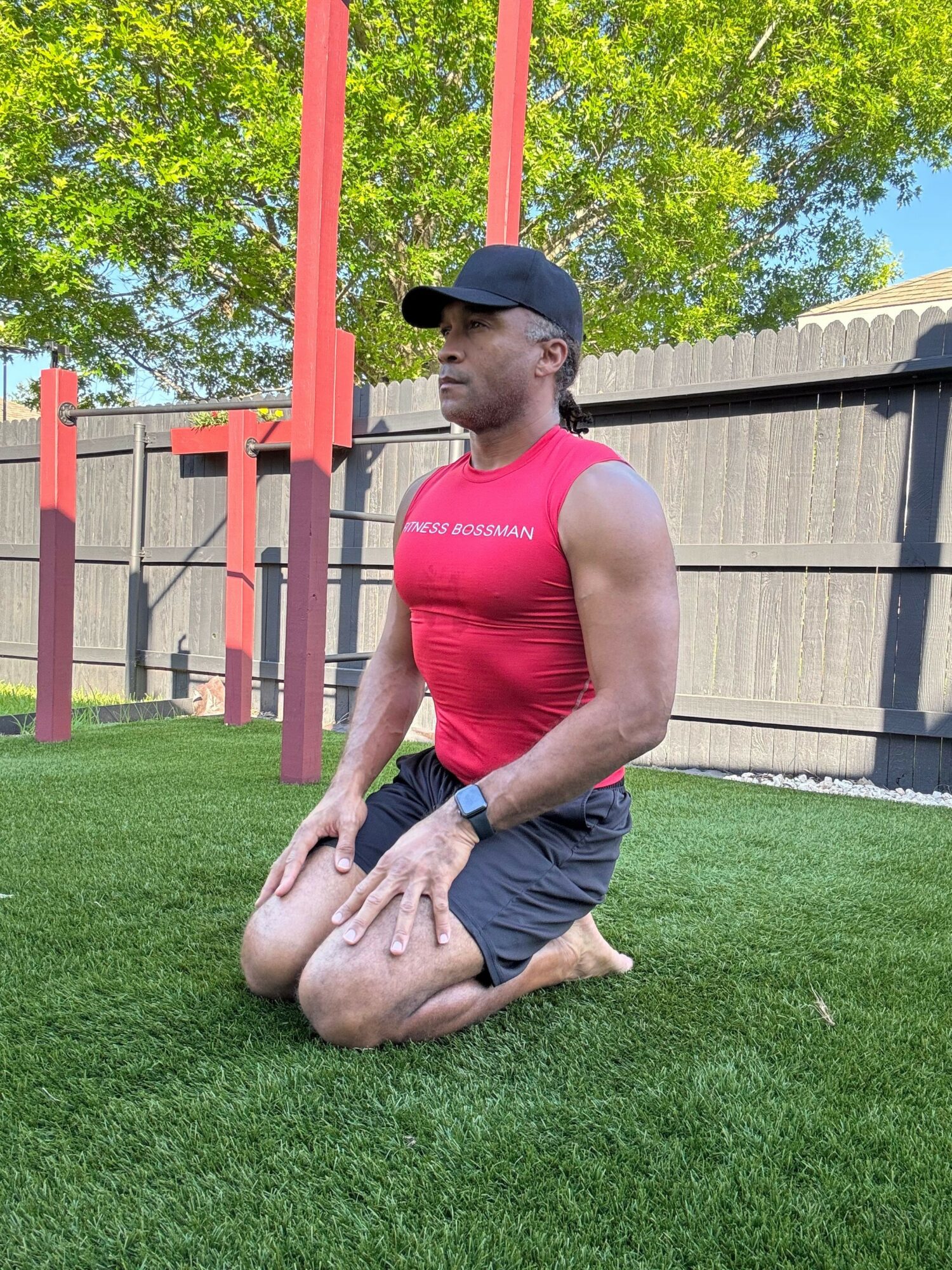 Person in red tank top and black shorts kneeling on green grass outdoors, with a wooden fence and trees in background.