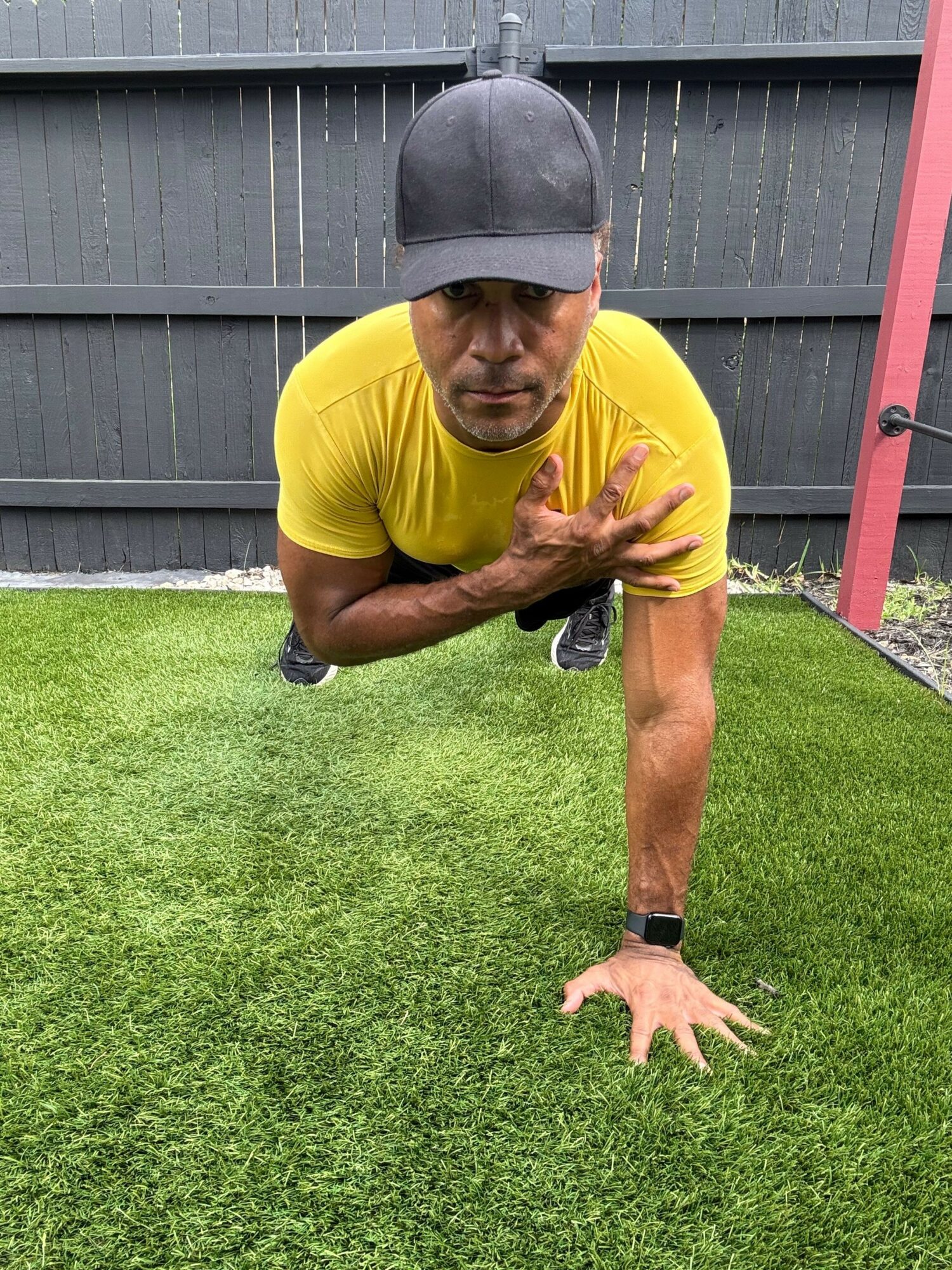 Person in yellow shirt and gray cap crouching on green grass, reaching forward with one hand, holding a water bottle.