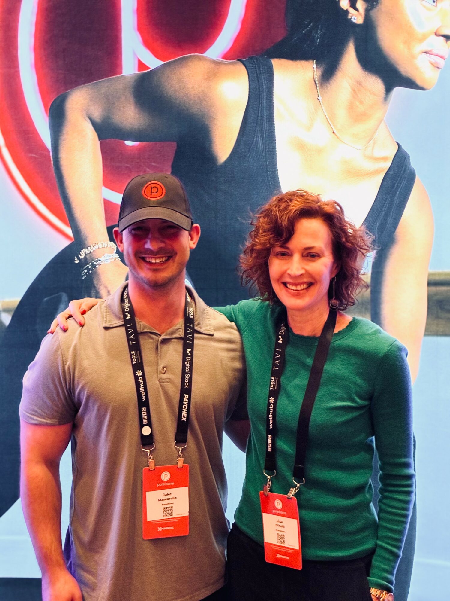 Two people smiling and standing together at an event, with a large poster of a woman in the background.