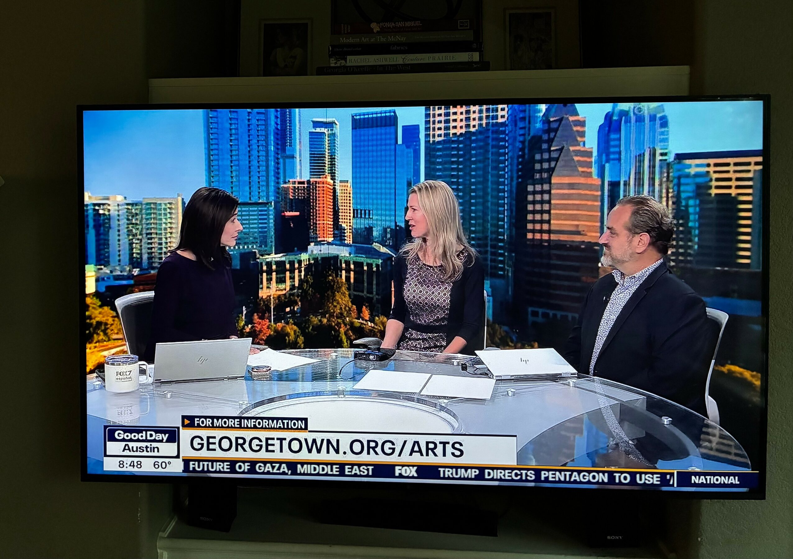 Three people sit at a news desk with a city skyline background, engaged in conversation, with papers and a laptop on the desk.