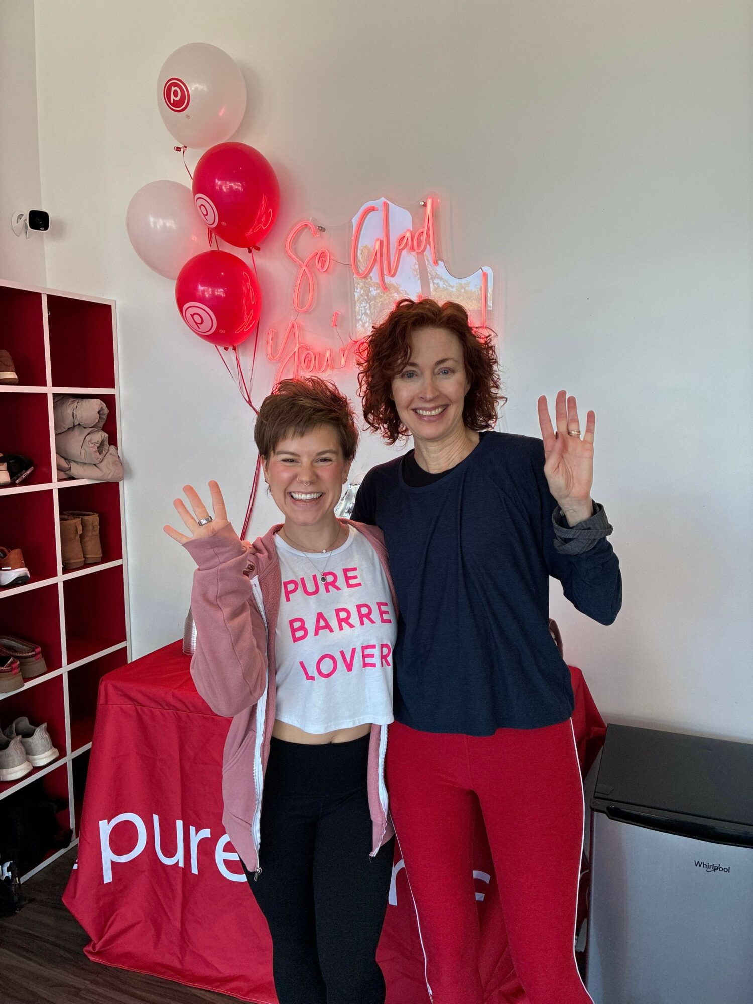 Two women standing in front of a white wall with red and white balloons, smiling and making peace signs.