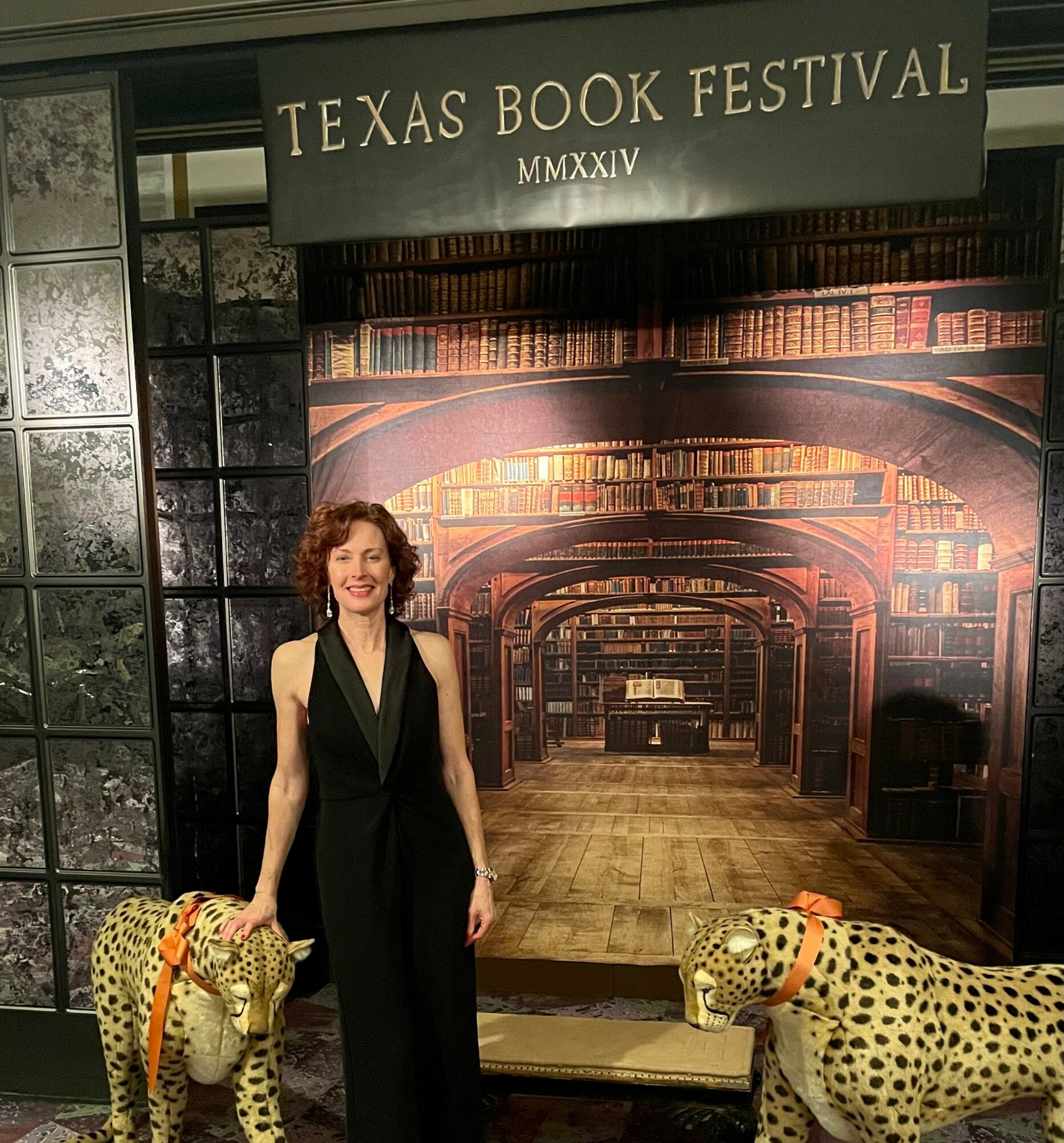 Woman in black jumpsuit standing between two stuffed cheetah toys in front of a library backdrop at Texas Book Festival.