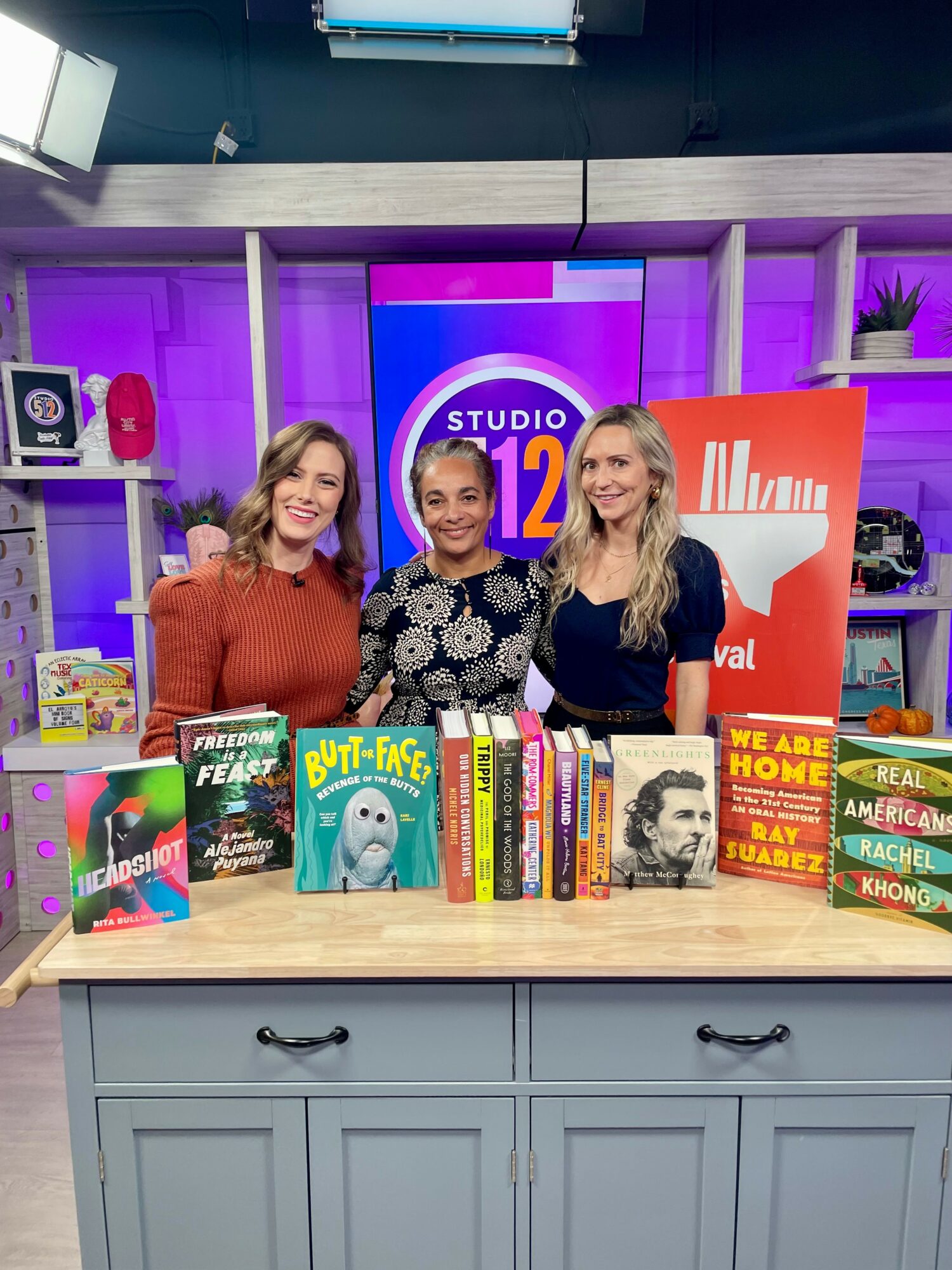Three women standing behind a table with colorful books, studio background with purple lighting, and a large screen.
