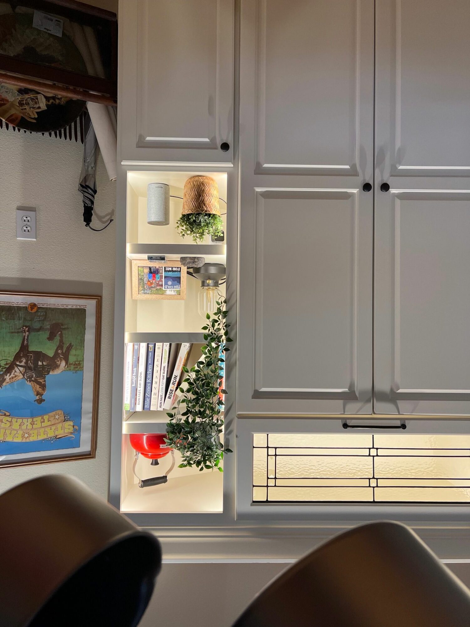 White cabinet with open shelves displaying plants, books, and decorative items, with a painting on the wall behind.