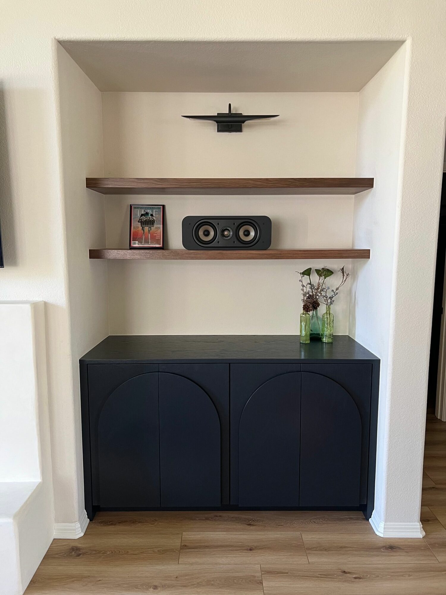Living room corner with wall-mounted TV, two wooden shelves, a speaker, and a cabinet, with a wooden floor.