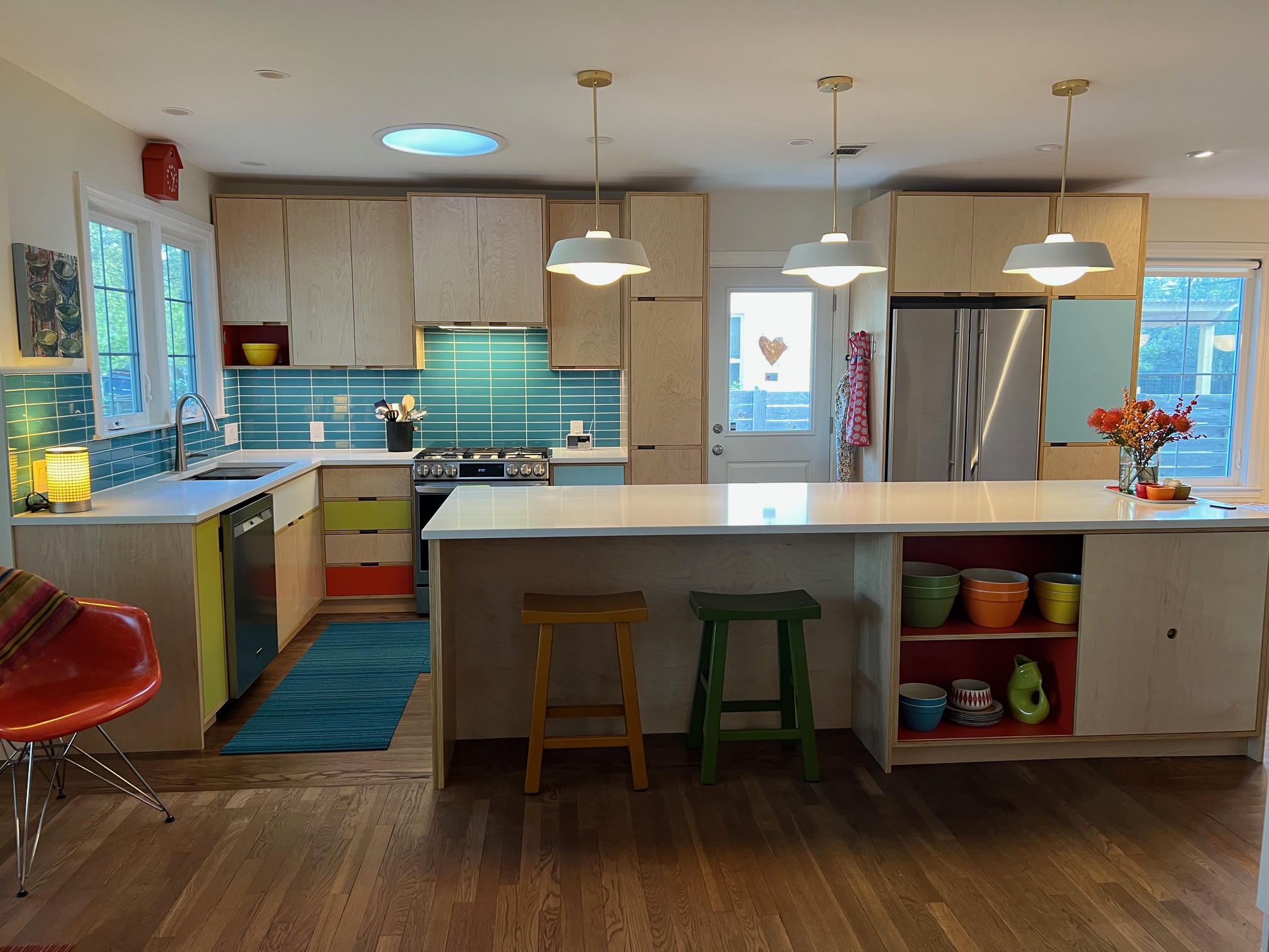 Modern kitchen with white cabinets, turquoise backsplash, island with stools, and pendant lights, with a wooden floor and window views.