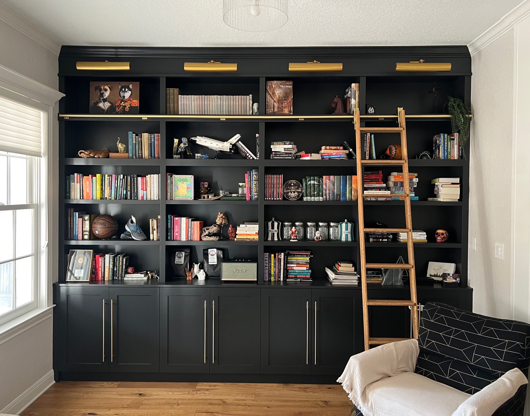Large black bookshelf filled with books, decorative objects, and a ladder leaning against it in a room with a window and a sofa.