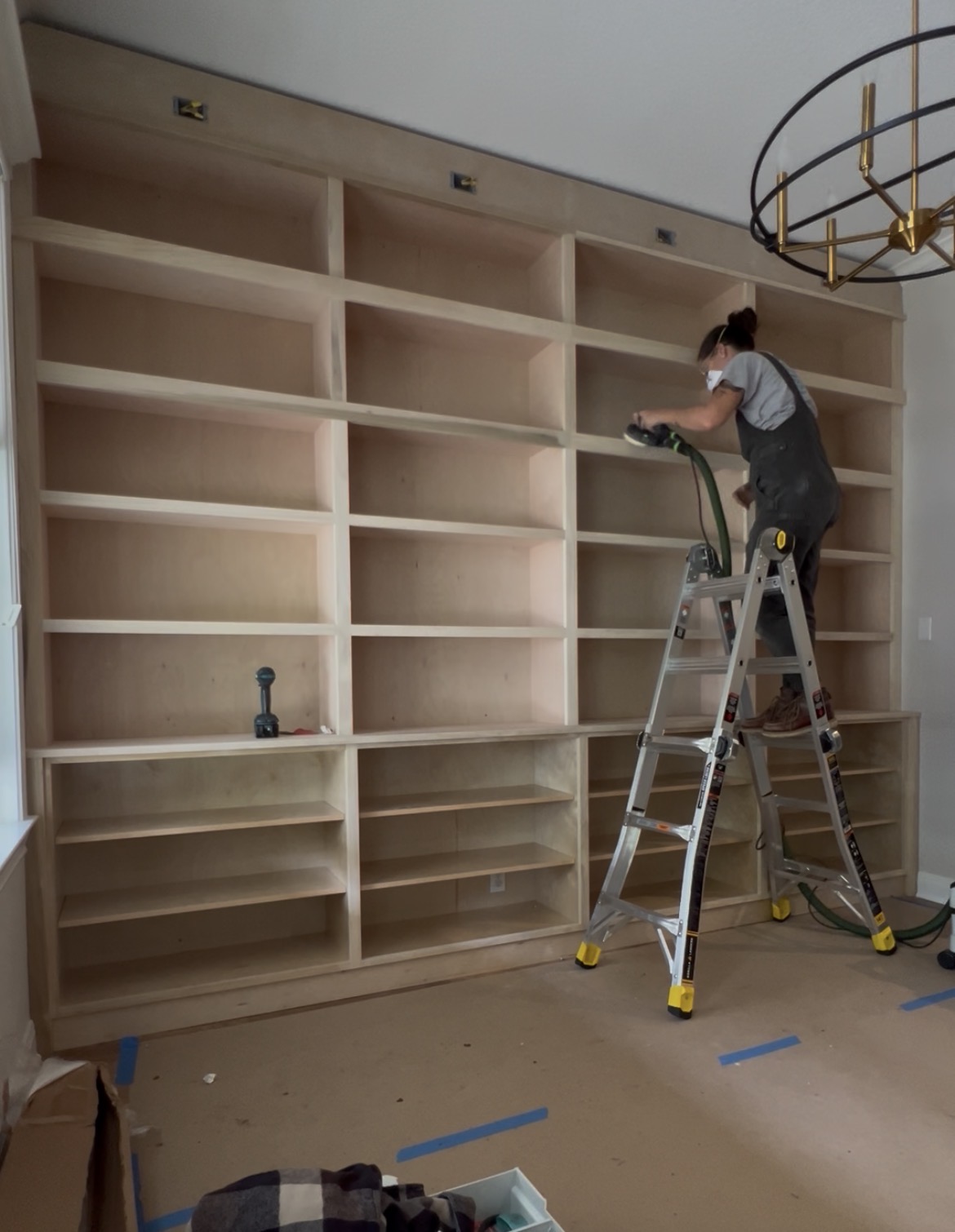 Person standing on a ladder installing or working on a large empty wooden bookshelf in a room with a ceiling light fixture.